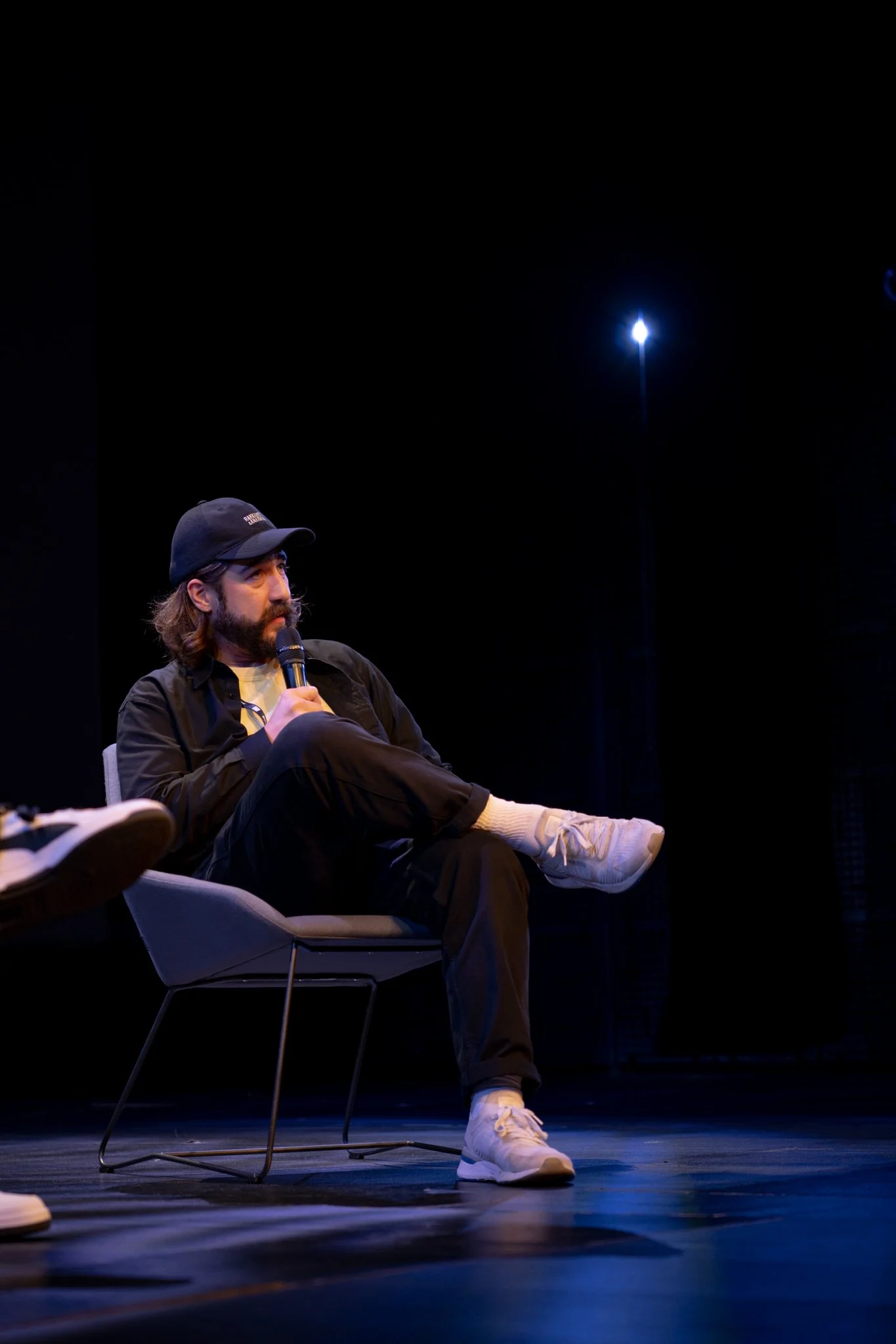 A man with a beard and long hair wearing a black cap, black jacket, and white sneakers, sitting on a chair on a stage, holding a microphone and speaking.