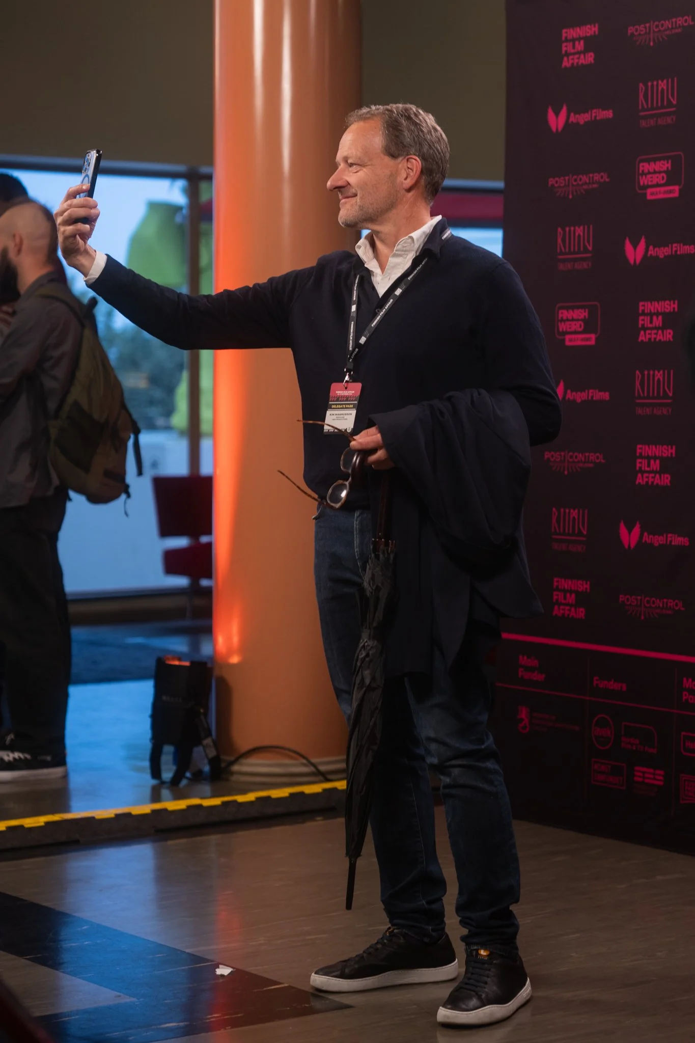 Man taking a selfie with his smartphone at a film event, standing in front of a backdrop with logos and text.