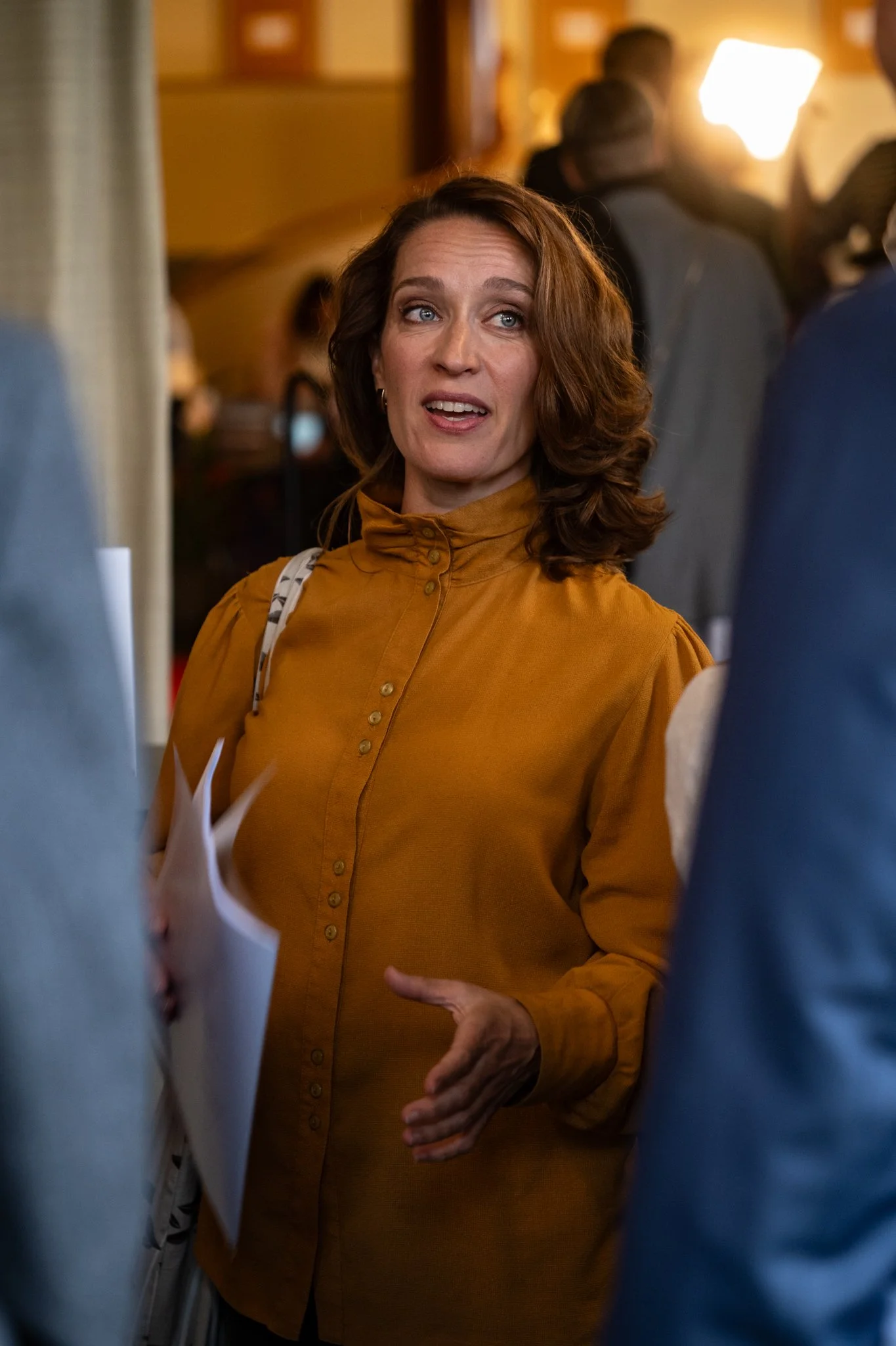 A woman with shoulder-length brown hair, wearing a mustard-colored blouse, appears to be speaking and gesturing with her left hand in a crowded indoor setting with warm lighting.