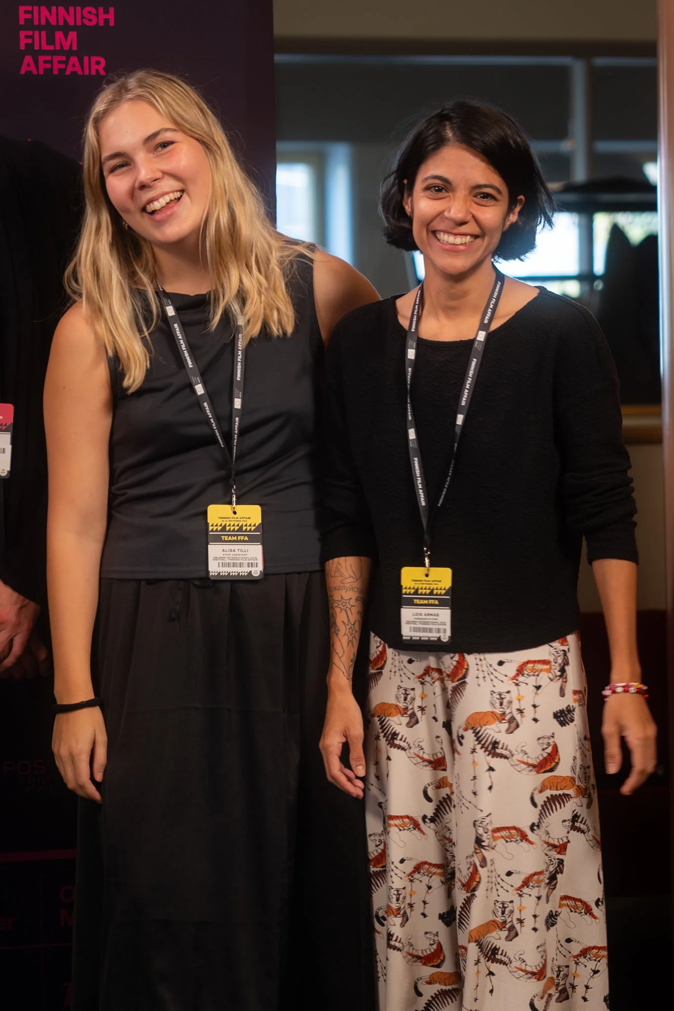 Two women standing together at an event, both wearing lanyards with badges, smiling for the photo. The woman on the left has long blonde hair, a black sleeveless top, and a black skirt. The woman on the right has short black hair, a black sweater, an