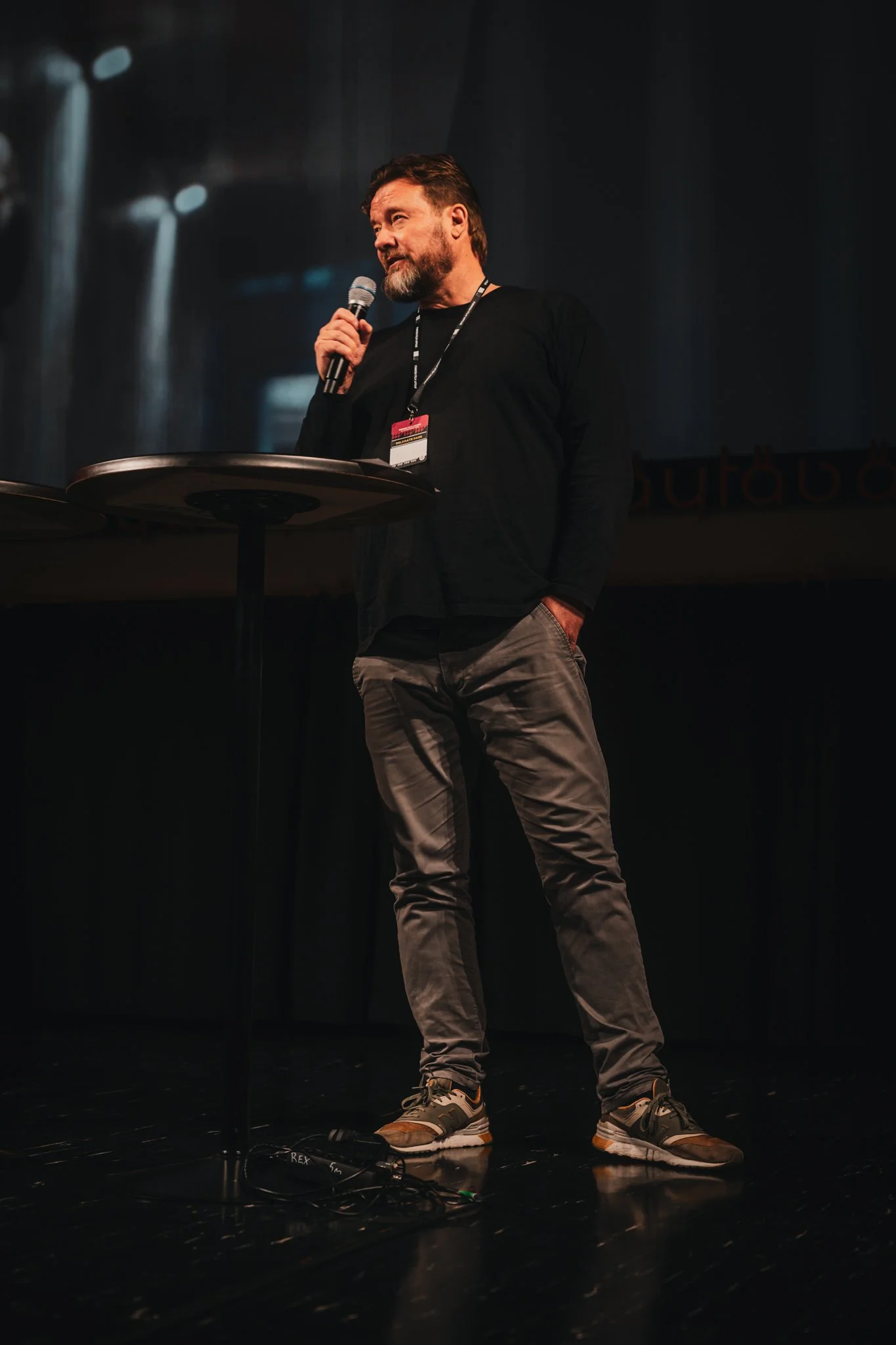 A man standing on a stage, holding a microphone in his right hand, wearing a black long-sleeve shirt, gray pants, and sneakers, with a lanyard around his neck, speaking or presenting to an audience.