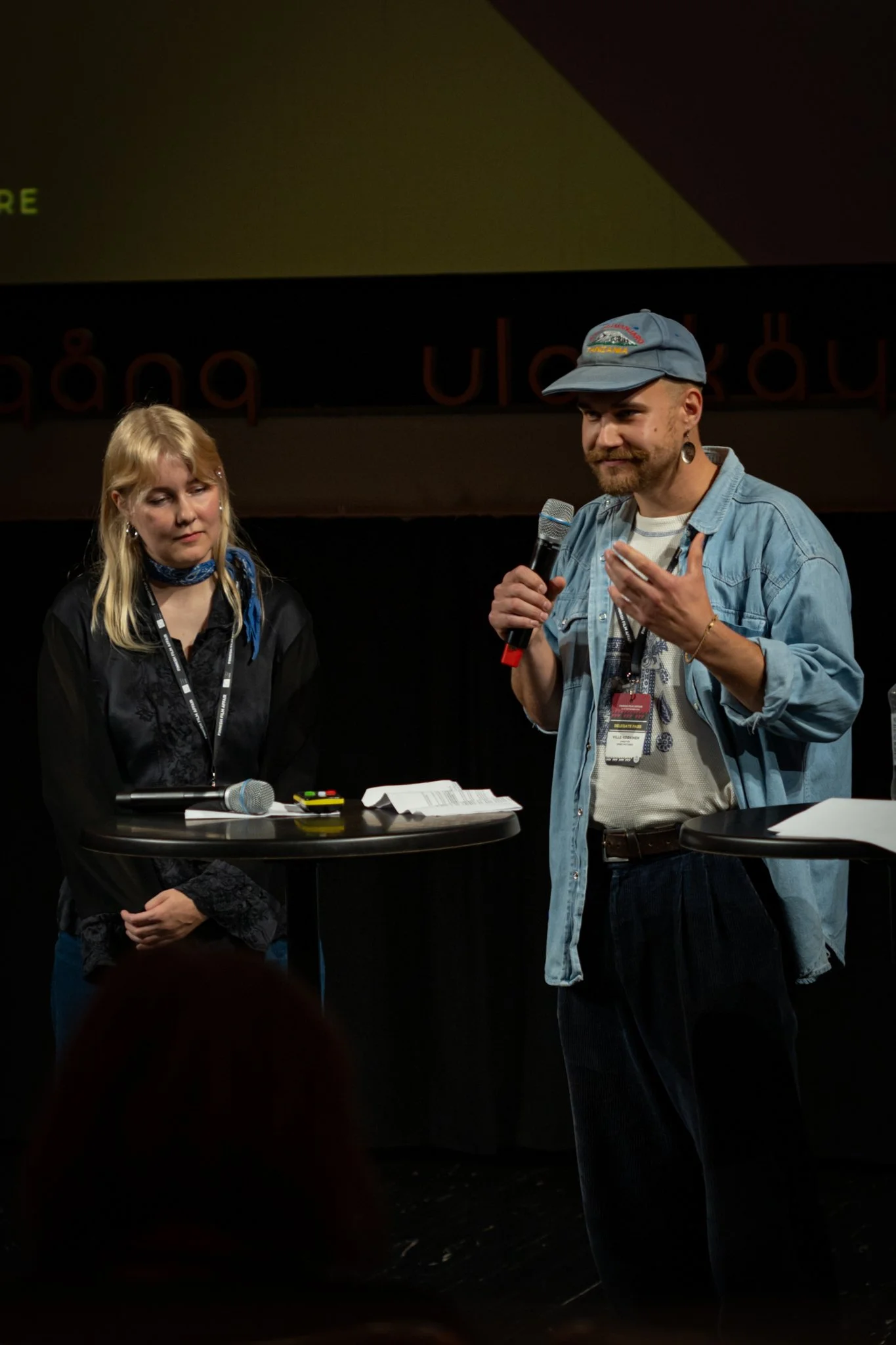 A man and a woman are standing at small high tables on a stage. The man is holding a microphone and speaking, while the woman looks down at the table. There are papers and a microphone on the woman’s table. The background features a dark curtain and 