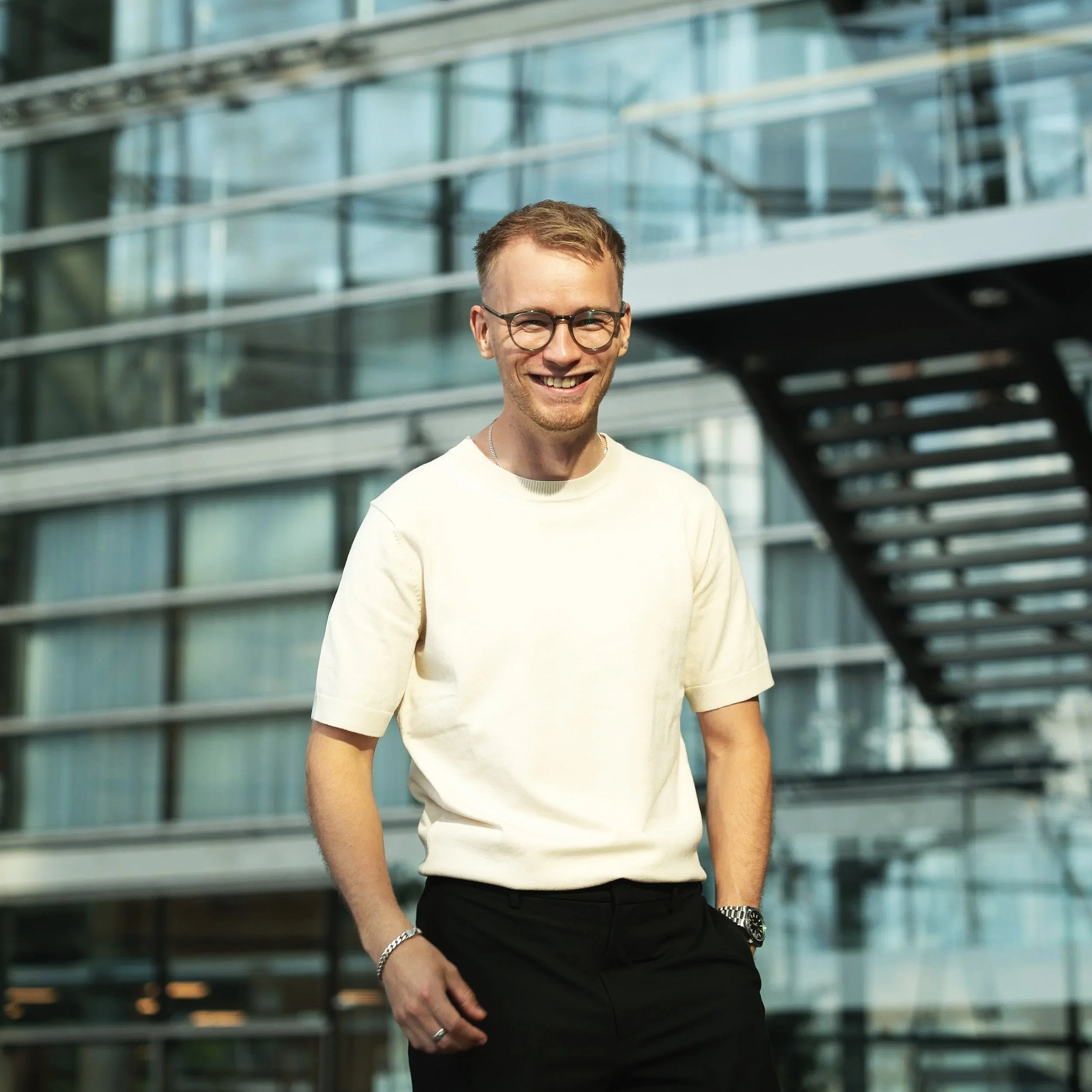Young man with glasses, short light brown hair, smiling, wearing a cream short-sleeve shirt, dark pants, standing outdoors near modern glass building with stairway.