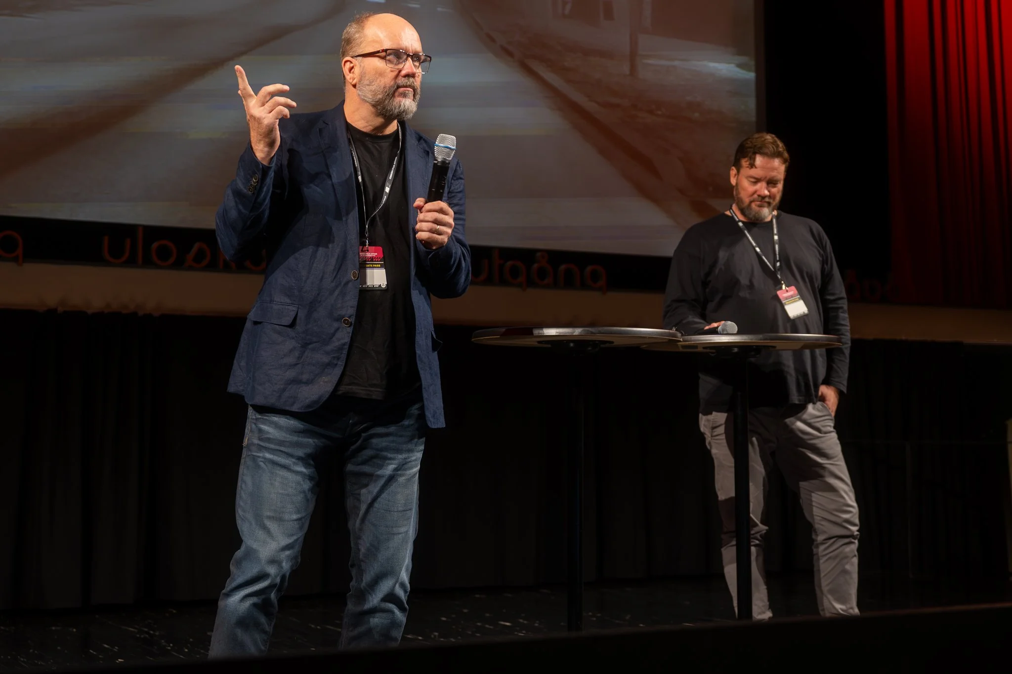 Two men standing on a stage with microphones; the man on the left is speaking while the man on the right listens, with a large screen behind them.
