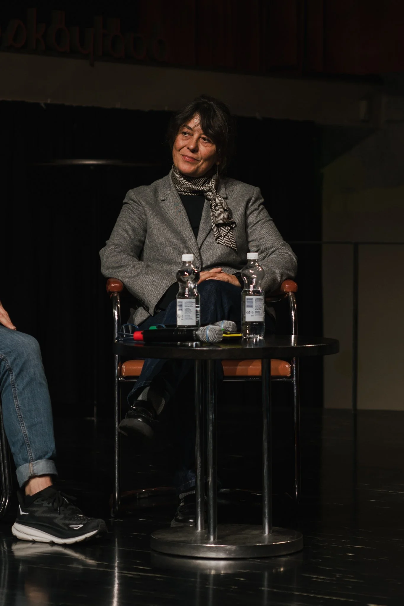 A woman in a gray blazer sitting at a small round table with two bottles of water, two microphones, and some electronic devices, on a dark stage.