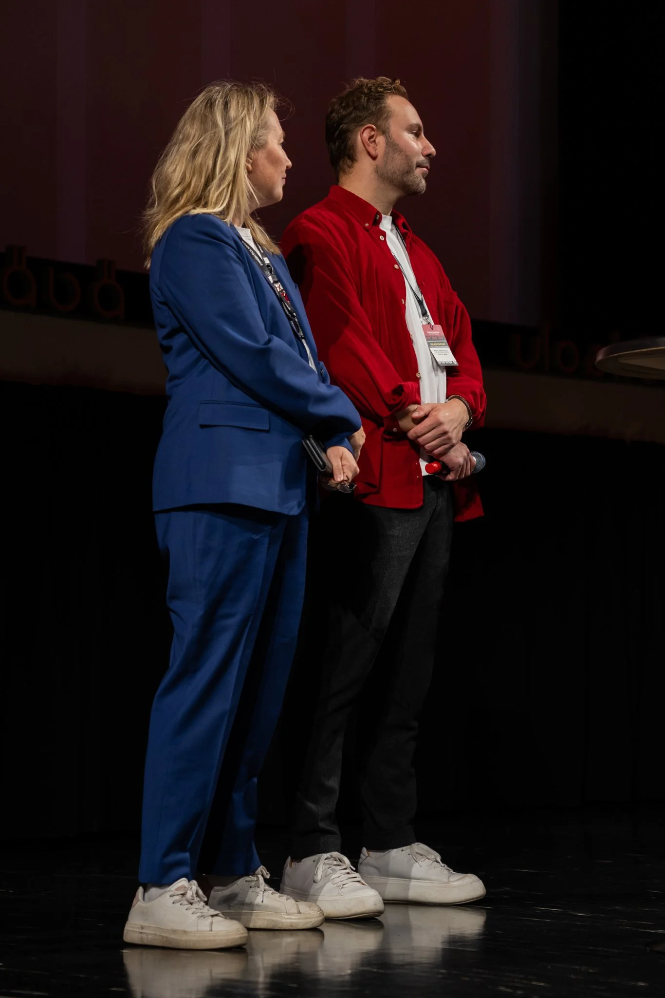 A woman in a blue suit and a man in a red jacket standing side by side on a stage, both holding microphones and wearing conference badges.