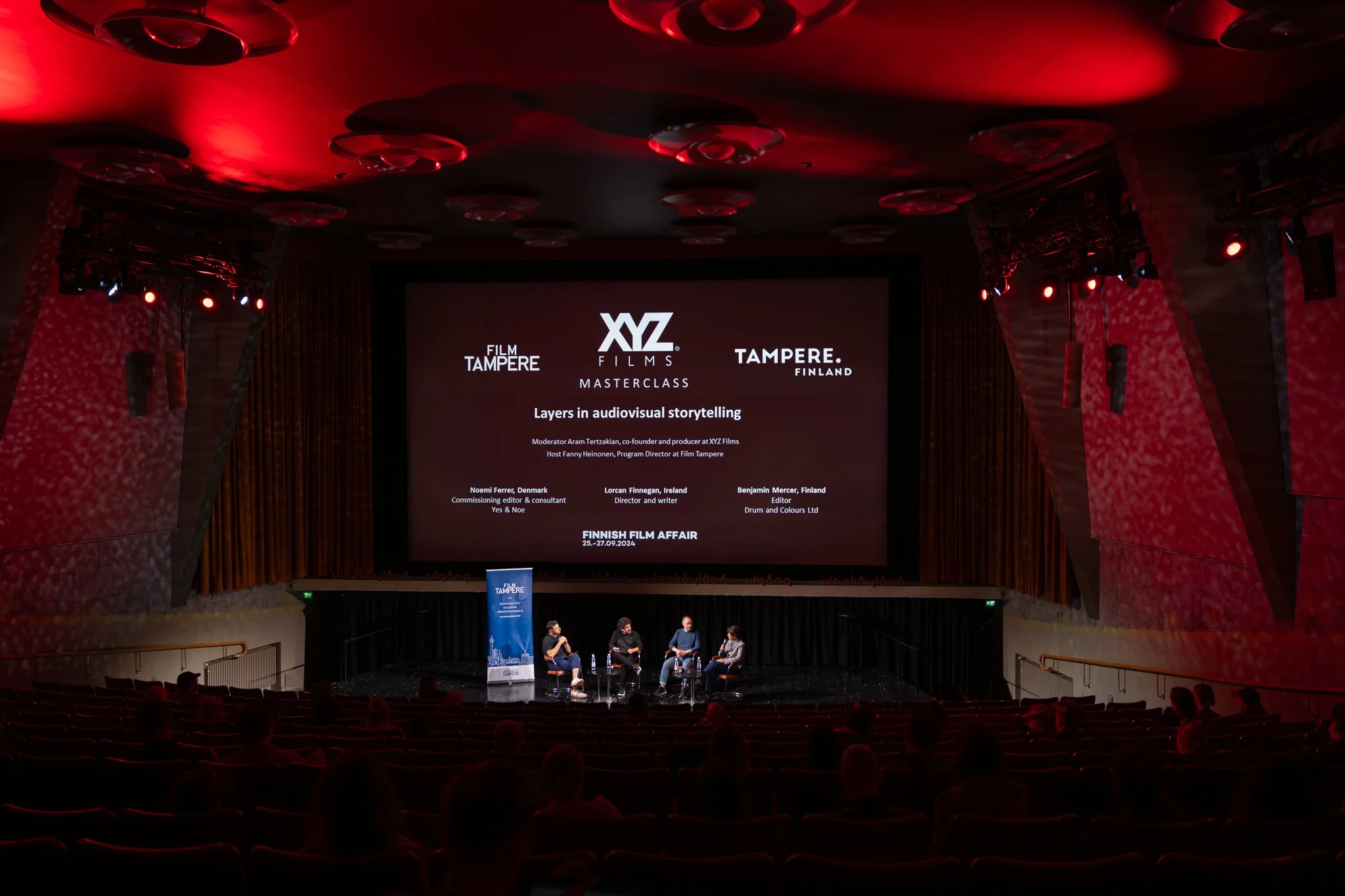 Panel discussion on a stage in a theatre or conference hall, with four speakers seated in chairs and a large screen behind them displaying the event details. The room has red and black lighting and an audience seated in front.
