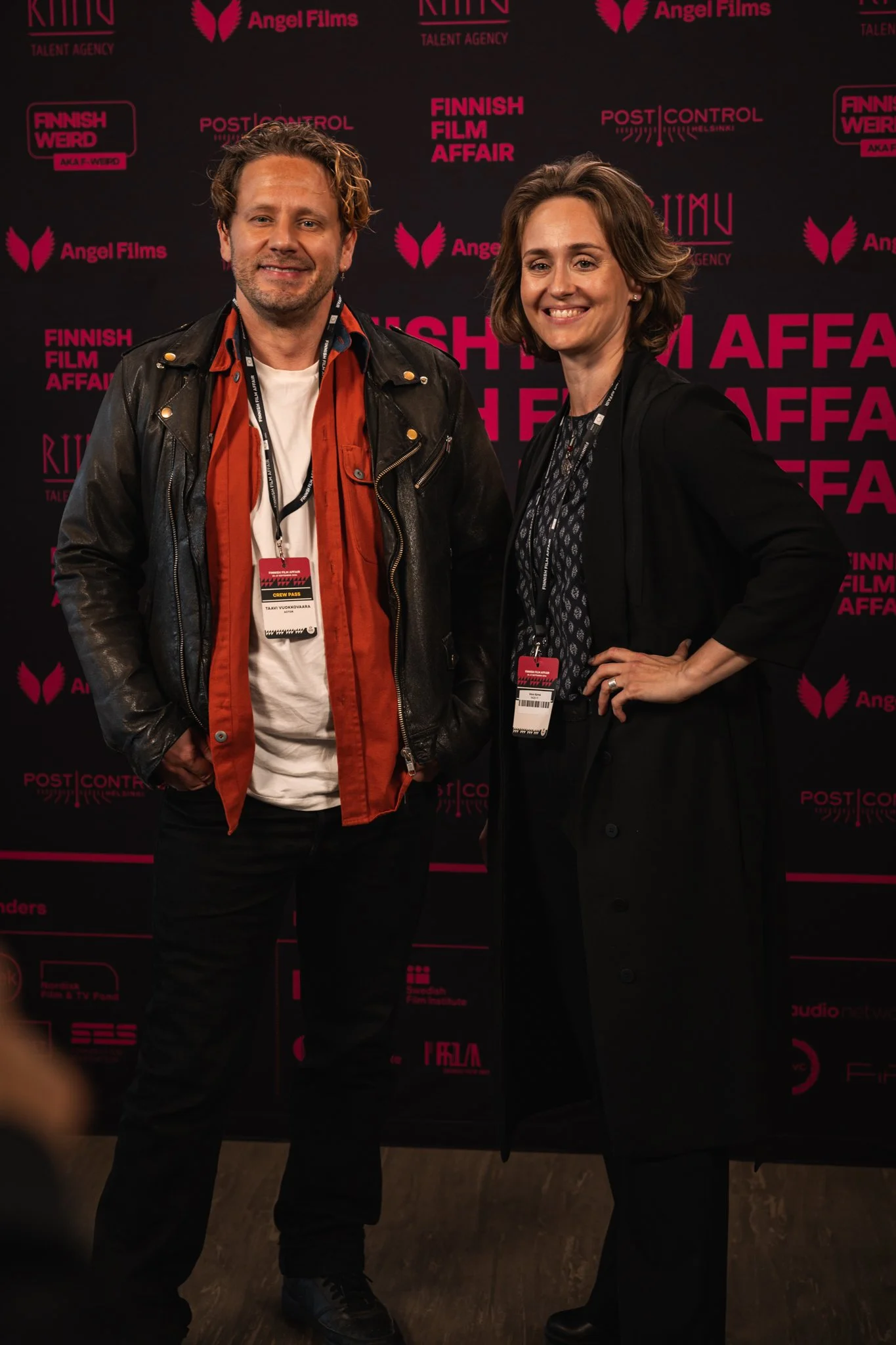 Two people standing in front of a black backdrop with pink text and logos related to Finnnish film and film industry, posing for a photo at an event.
