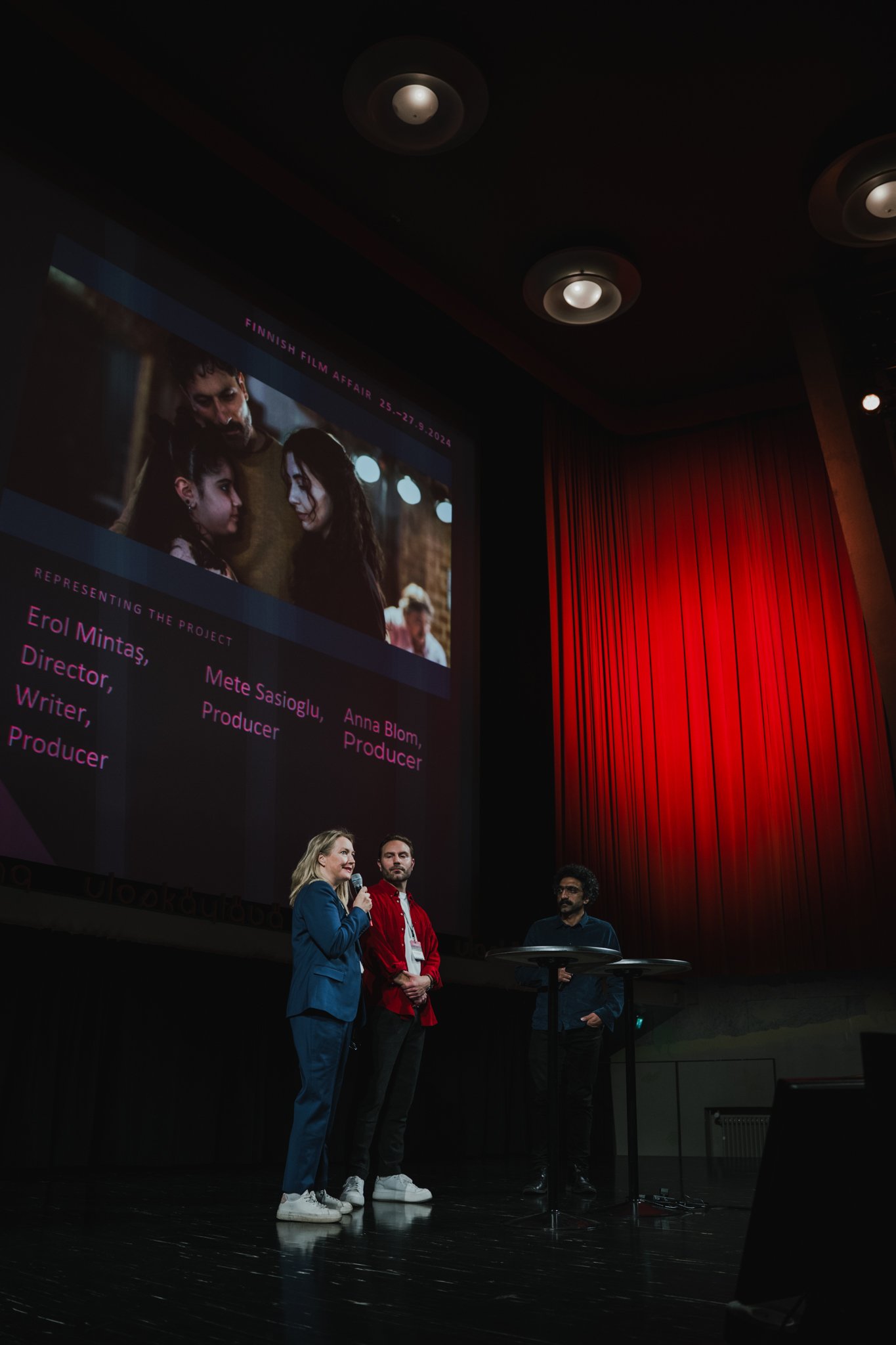 People on stage at a film event, with a large screen behind them displaying a film poster or title, and individuals speaking into a microphone.