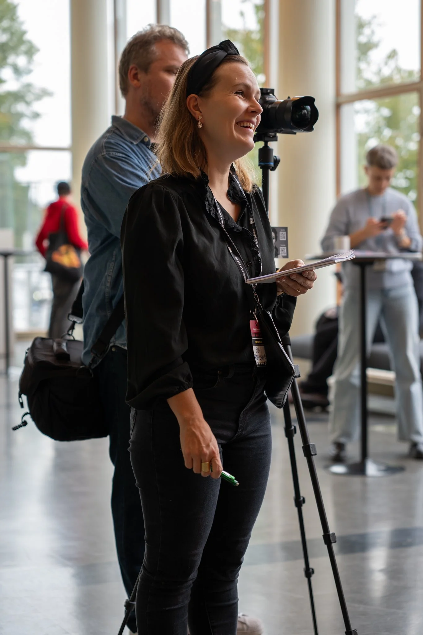 A woman smiling while operating a camera on a tripod in a bright indoor space, with a man standing behind her and a person using a phone in the background.