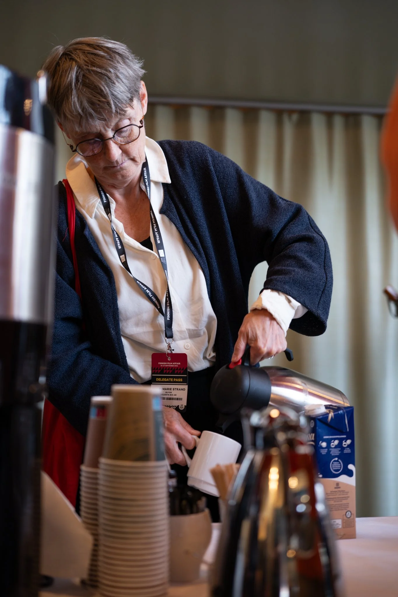 A woman with short hair, glasses, and a black cardigan preparing coffee using a coffee pot at a buffet or conference setting.