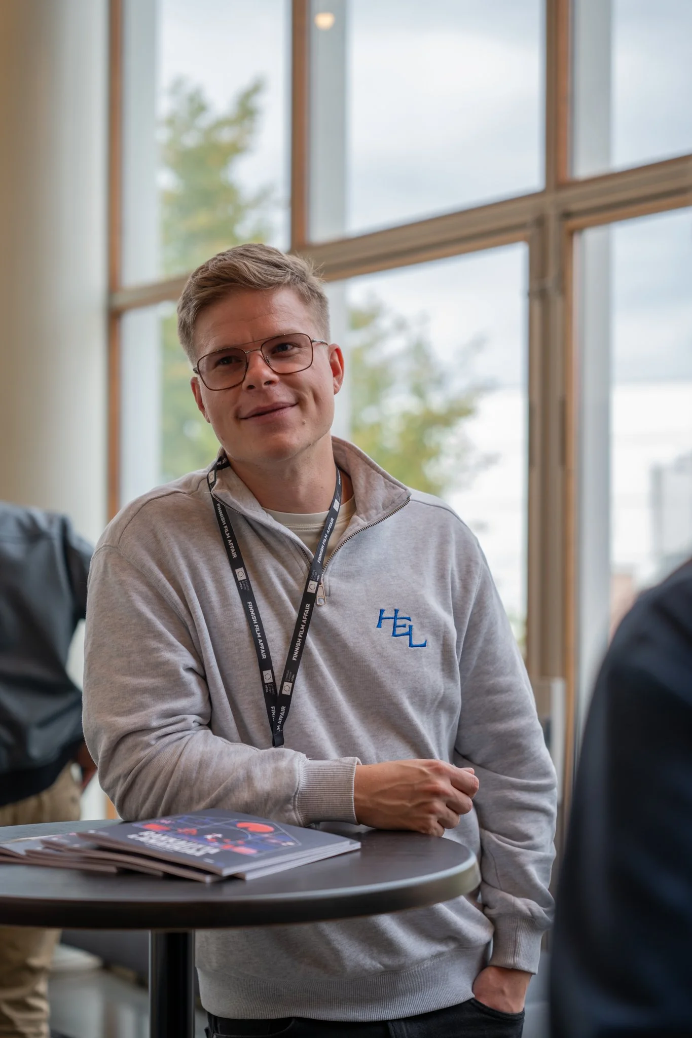 A young man with glasses and a light gray pullover sitting at a table, smiling indoors with large windows behind him.