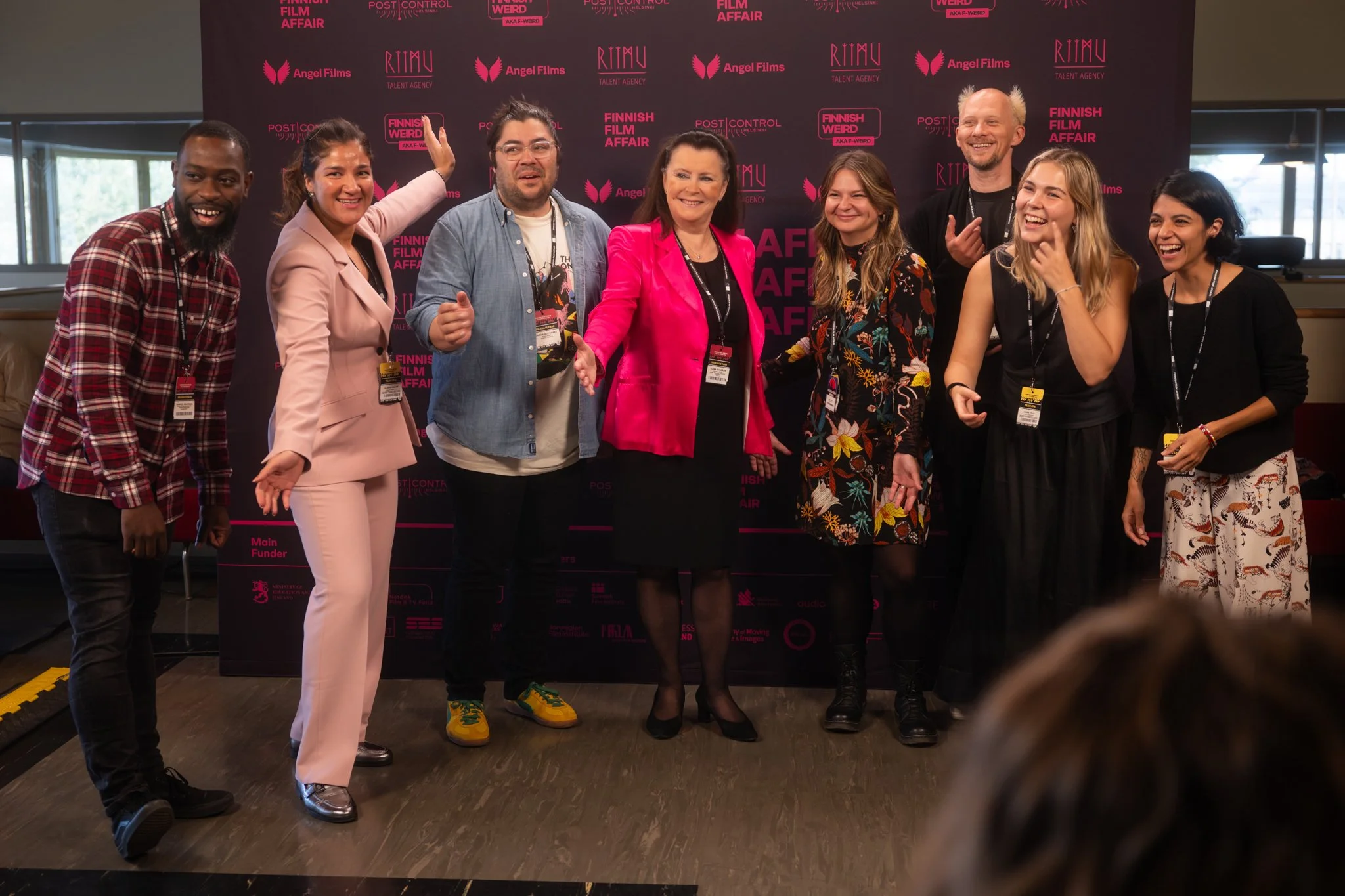 Group of nine diverse people posing together at a film event, standing in front of a backdrop with various logos and pink text, smiling and interacting.