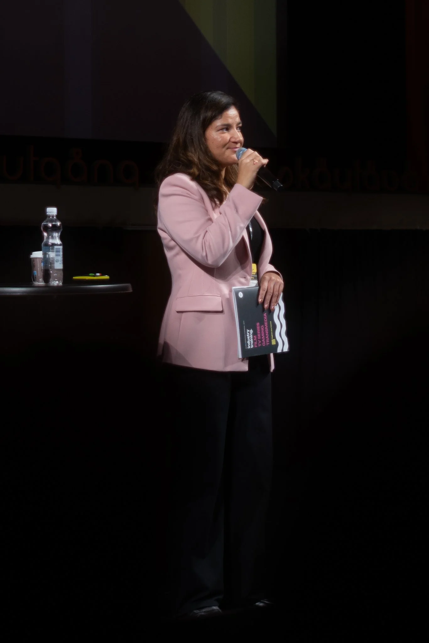 Woman in pink blazer speaking into a microphone on stage with water bottle and booklet