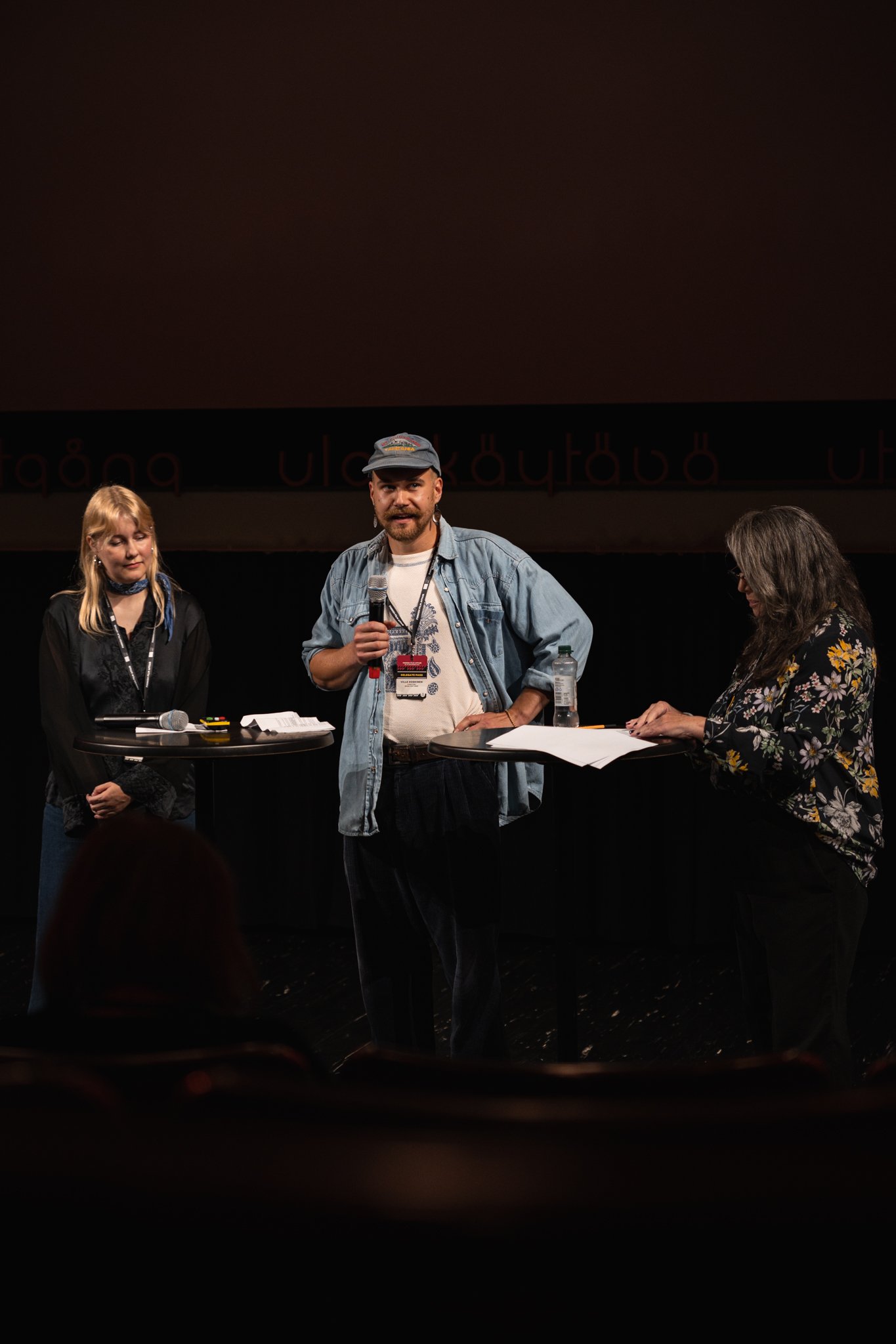 Three people standing on a stage with microphones and papers, participating in a discussion or presentation, with a dark backdrop and audience in front.