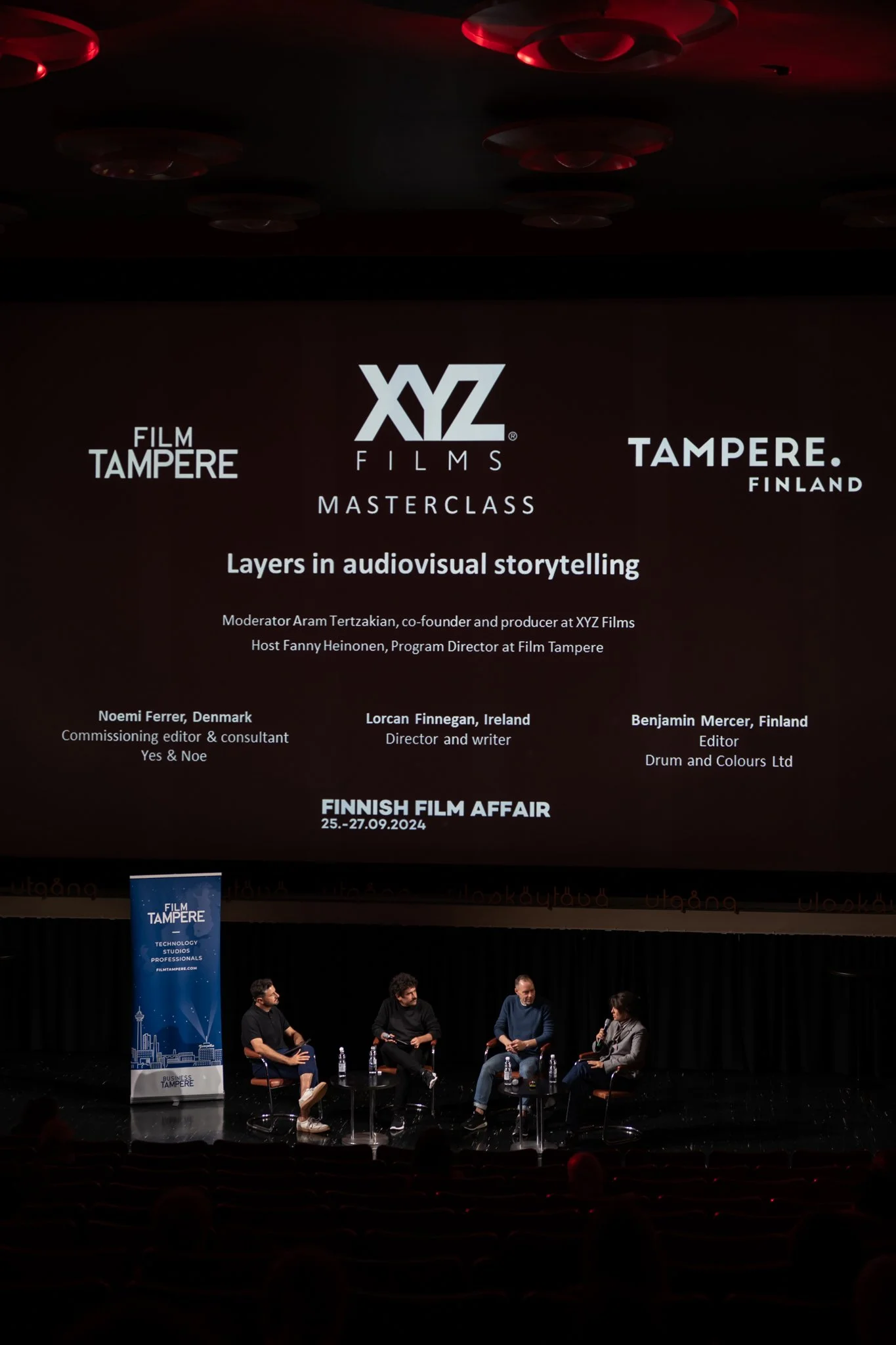 Panel discussion at Finnish Film Affair 2024, with four people seated on stage, a large screen displaying event details, and a blue banner reading 'Film Tampere' on the left.
