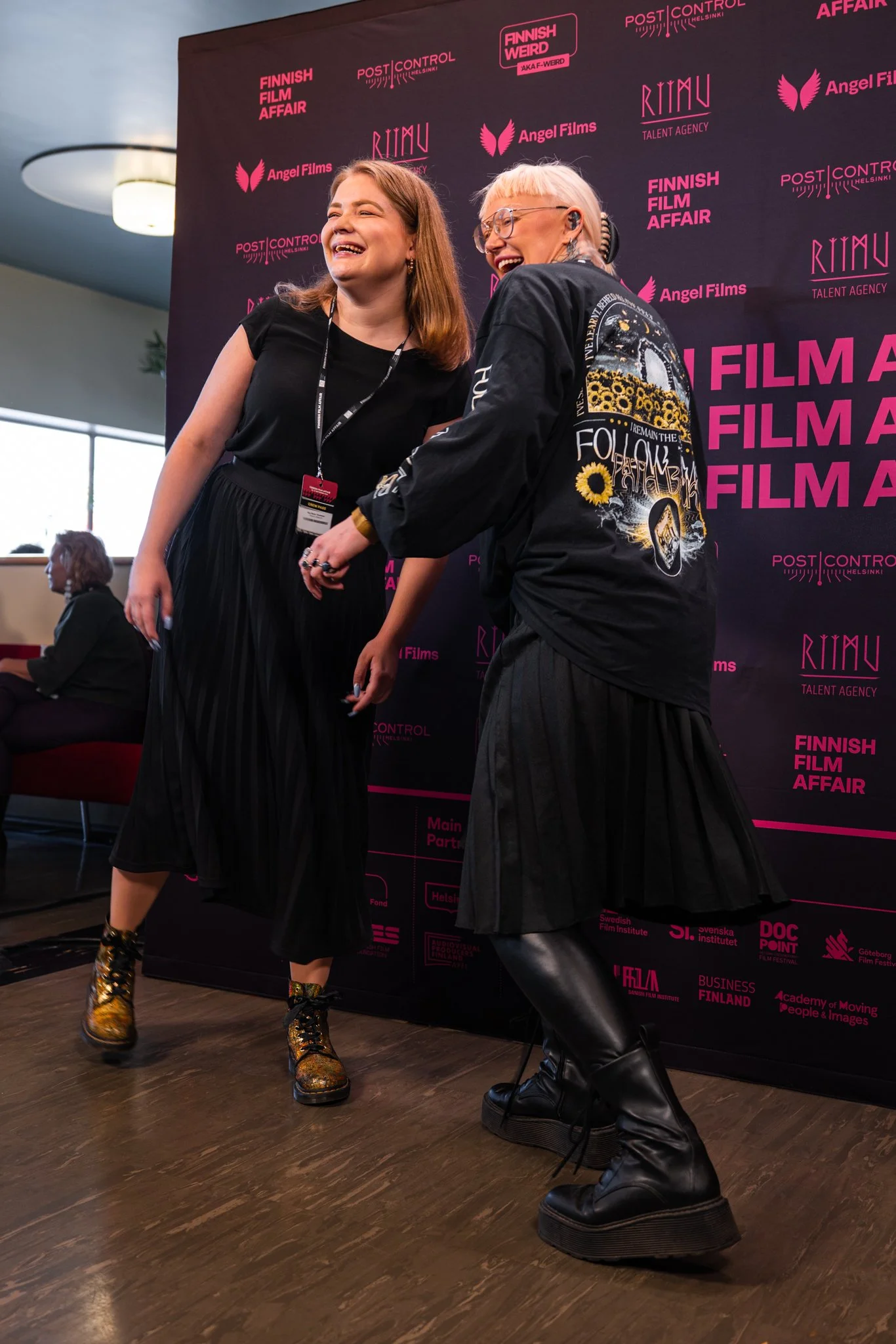 Two women laughing and holding hands on a stage at the Finnish Film Affair event, with a dark backdrop featuring pink text and logos, and a woman sitting in the background.