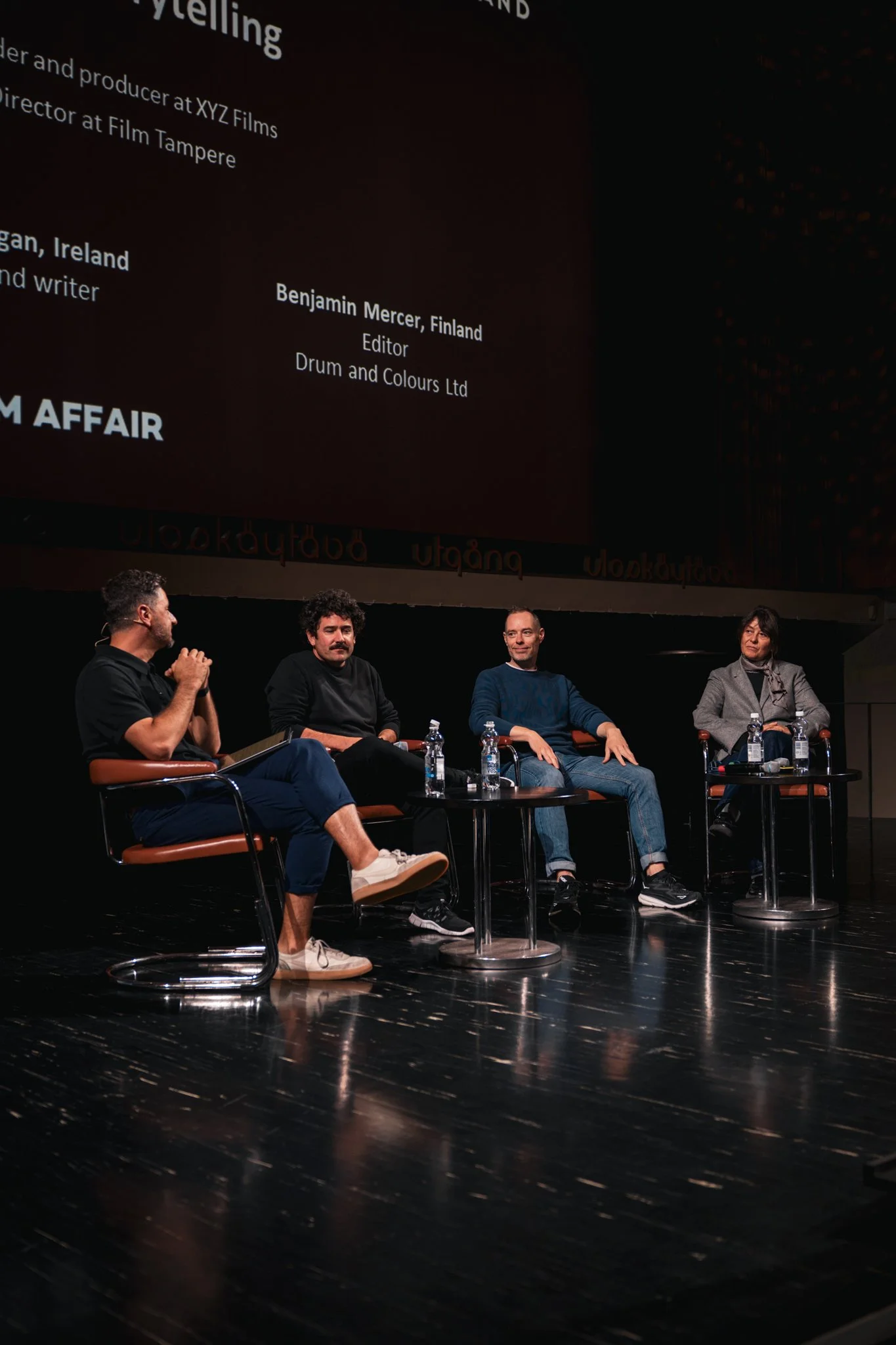 Four people sitting on a stage during a panel discussion, with water bottles on small tables in front of them, and a large screen behind displaying text and credits.