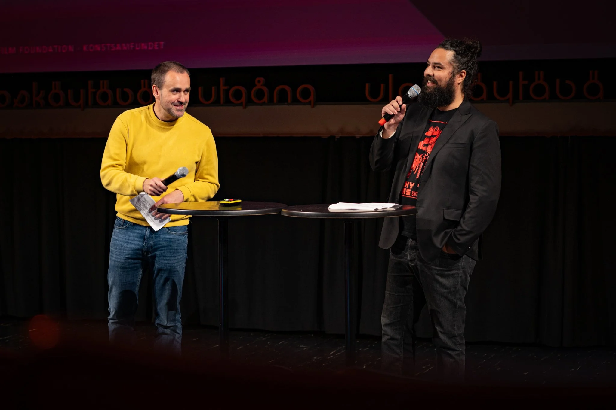 Two men on stage, one with a yellow sweatshirt holding a microphone and some papers, the other with a black blazer and graphic t-shirt, speaking into a microphone, with tables and a backdrop behind them.