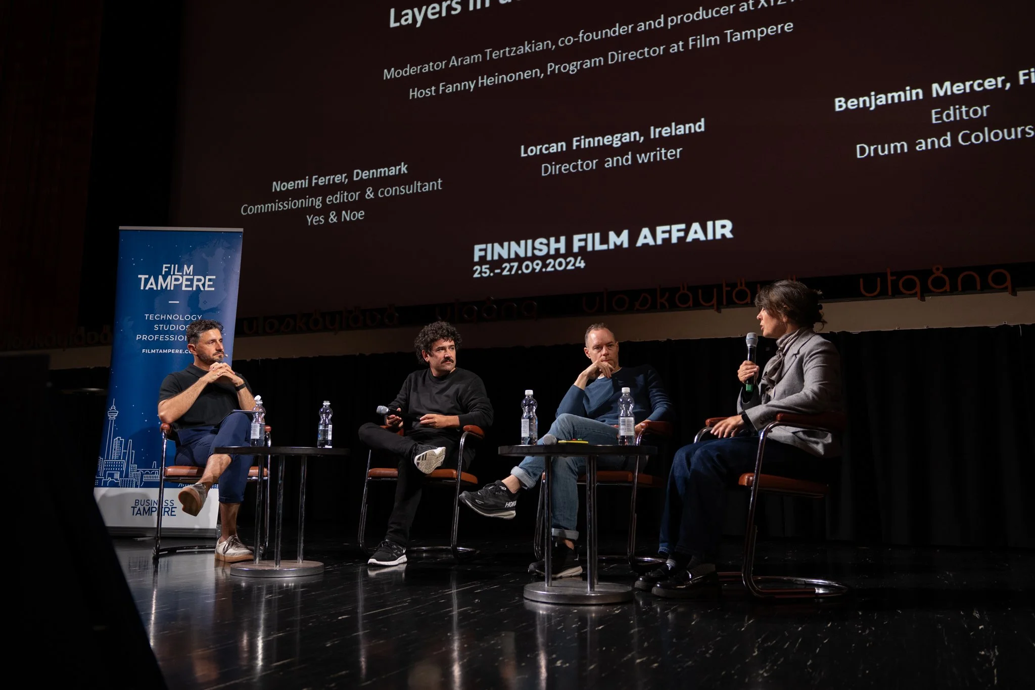 Panel discussion at Finnish Film Affair, with four individuals seated on stage with microphones and water bottles, backdrop displaying names and event details, and a blue banner on the left.