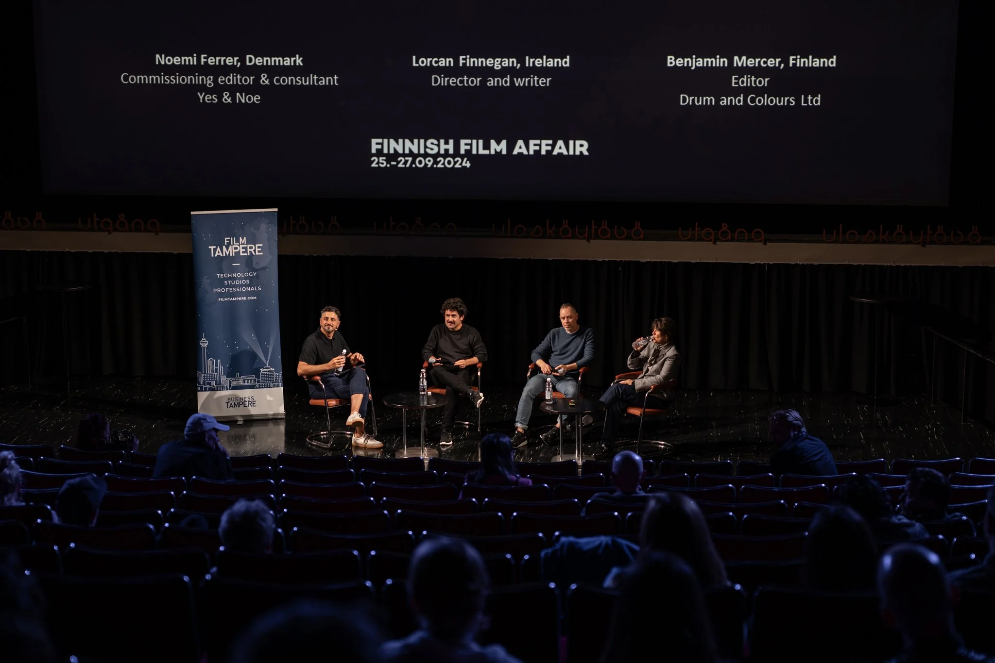 Panel discussion on stage at Finnish Film Affair event, with four speakers seated with water bottles, in front of a large screen displaying their names and titles, and a Finnish Film Affair banner on the side.