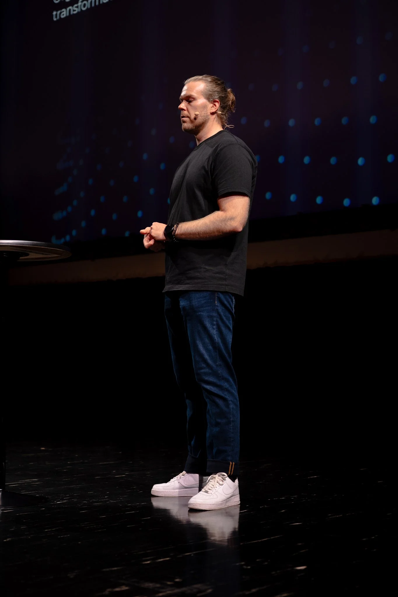 A man with long hair tied back, dressed in a black t-shirt, jeans, and white sneakers, standing on a stage during a presentation.