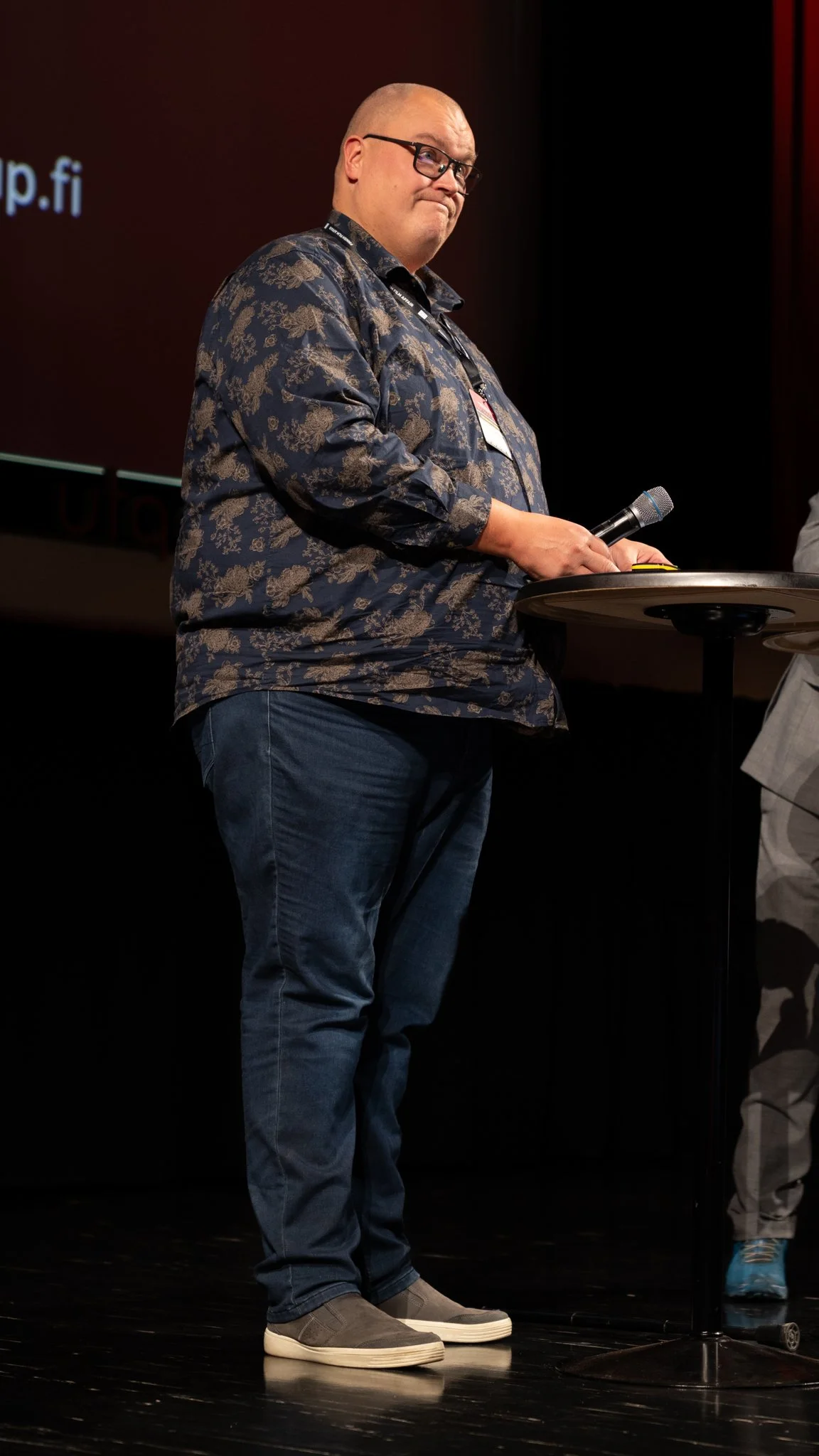 A man standing on a stage, holding a microphone and wearing glasses, a dark patterned shirt, and jeans, with a conference badge around his neck, during a presentation or event.