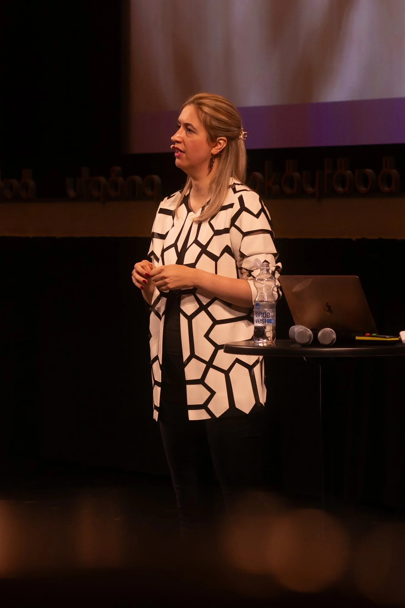 A woman with blonde hair wearing a black and white geometric patterned blazer, standing on stage in front of a large screen, with a table beside her holding a water bottle, two microphones, and a laptop.