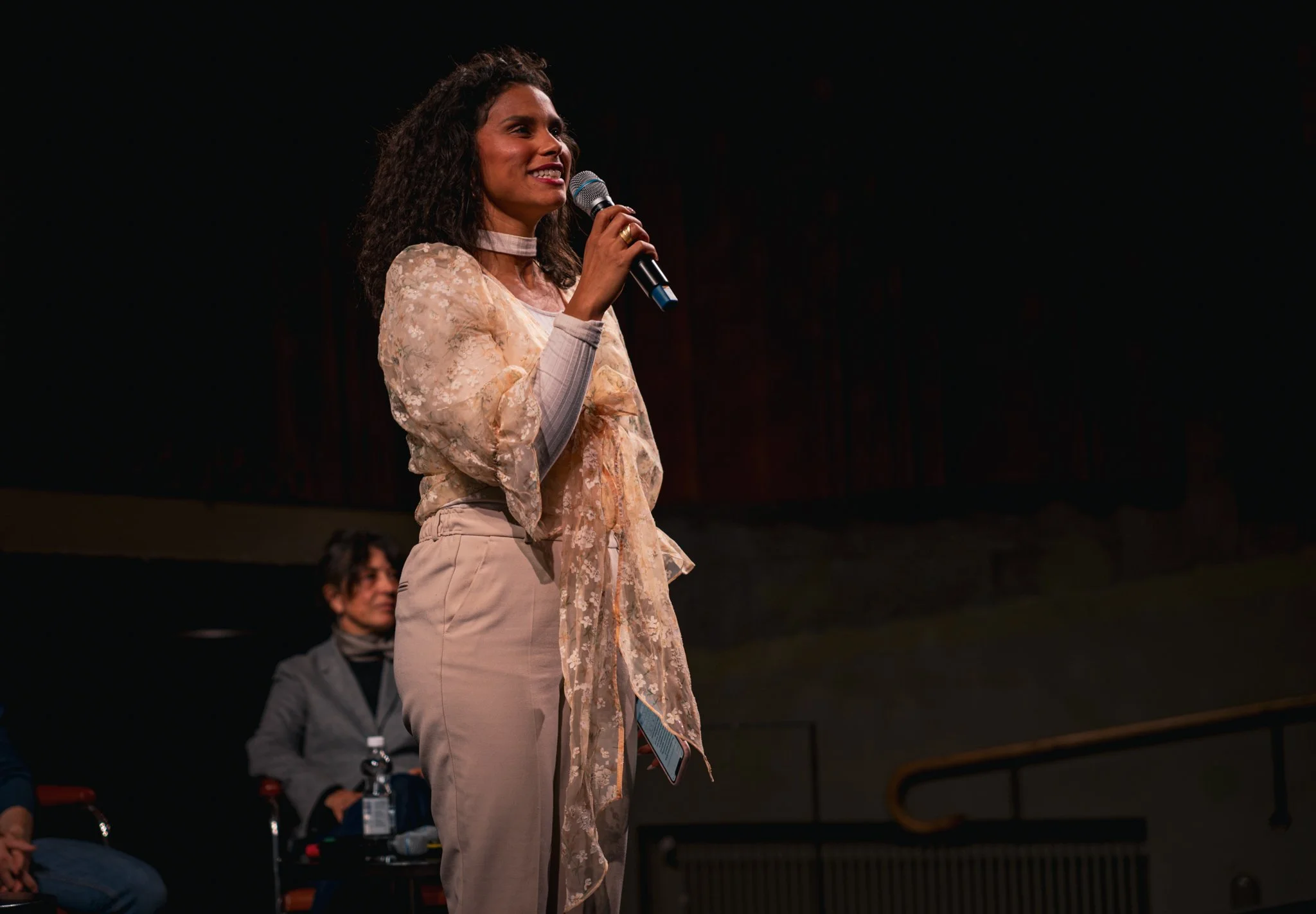 A woman speaking into a microphone on stage, wearing a floral blouse and beige trousers, with another woman sitting in the background.