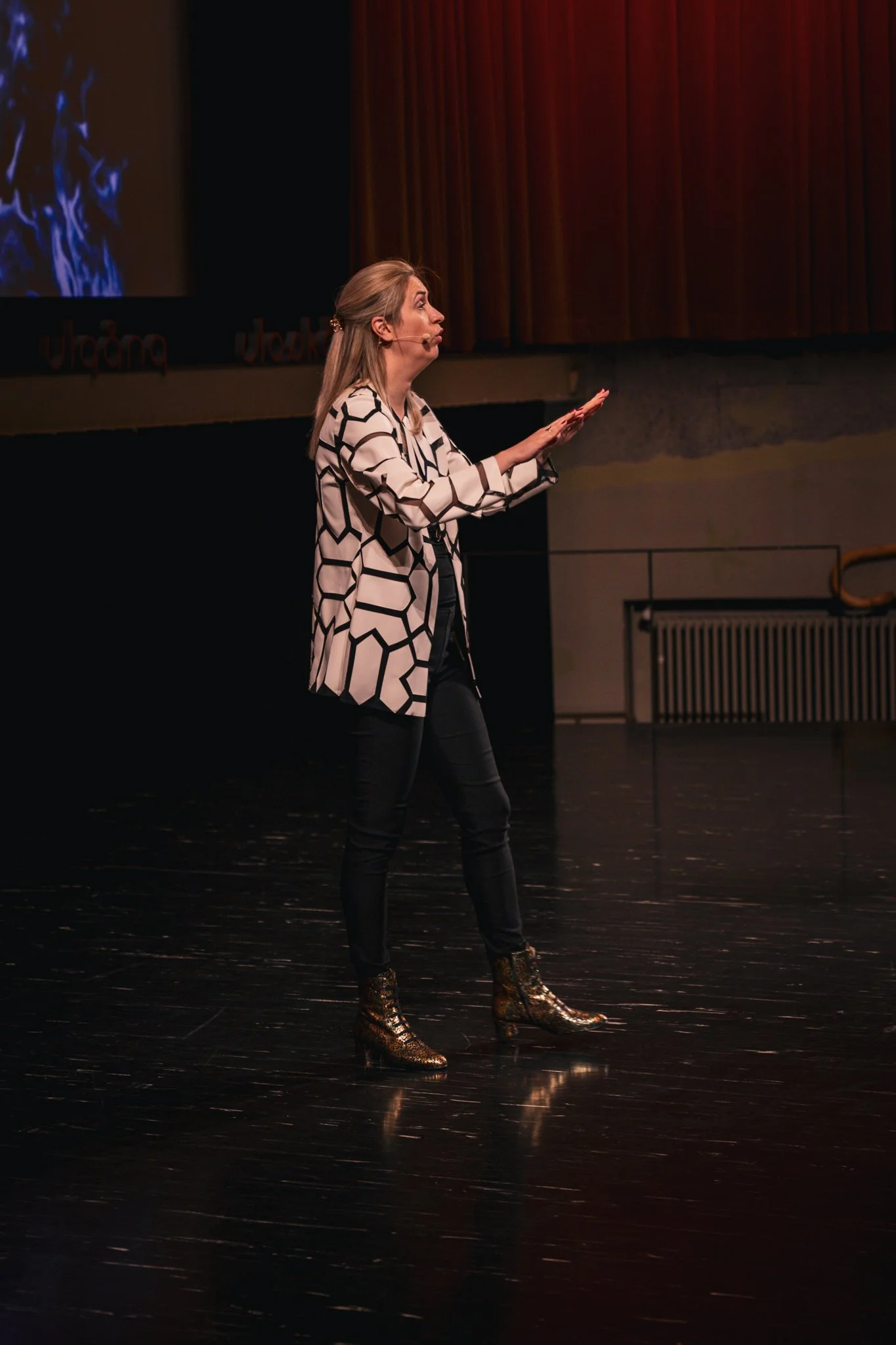 A woman on stage in a black and white patterned blazer, black pants, and gold snake print boots, speaking with her hands extended in front of her during a presentation or performance.