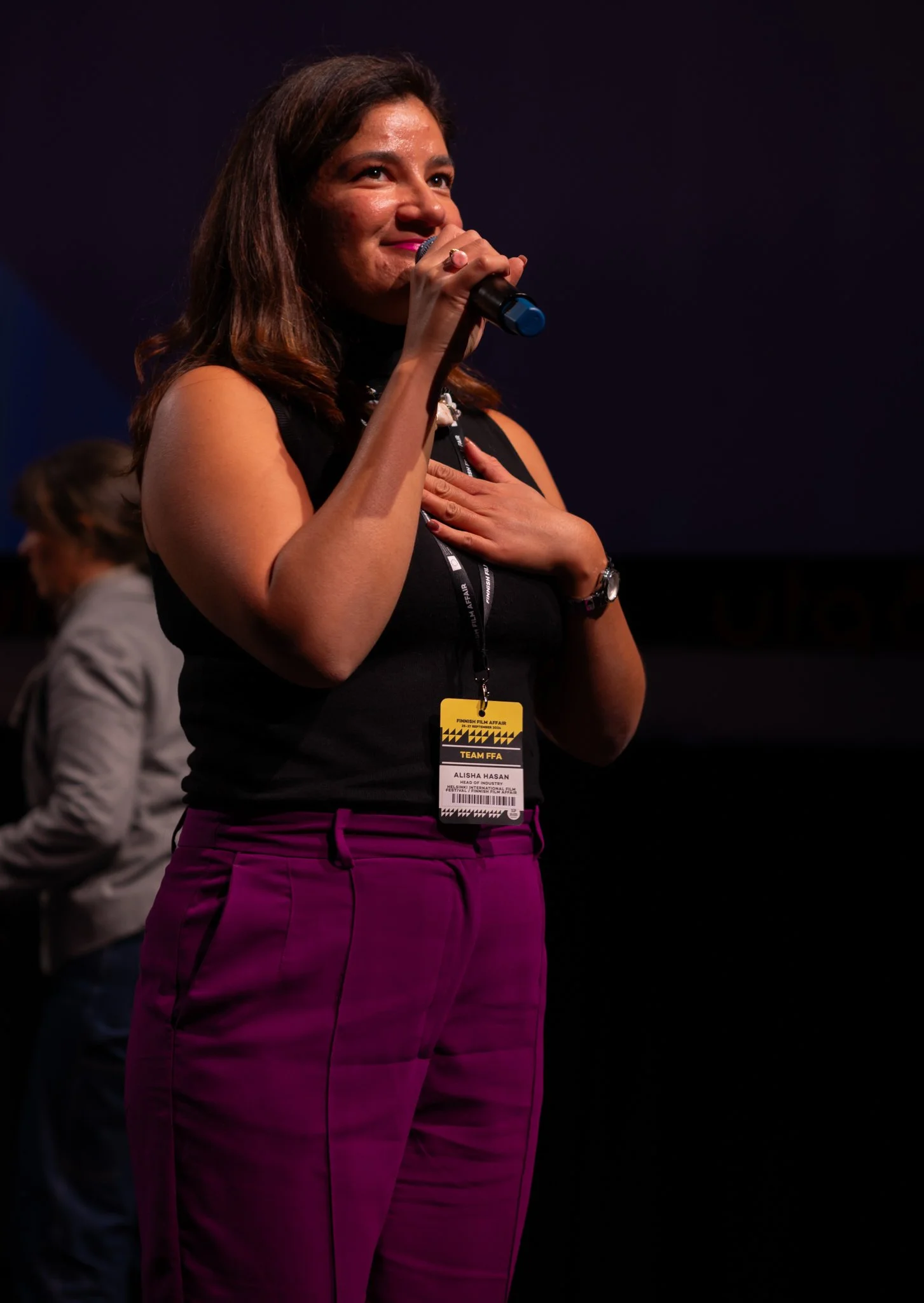 Woman on stage holding a microphone, wearing a black sleeveless top and purple pants, with a badge around her neck, hand on her chest, appearing emotional.
