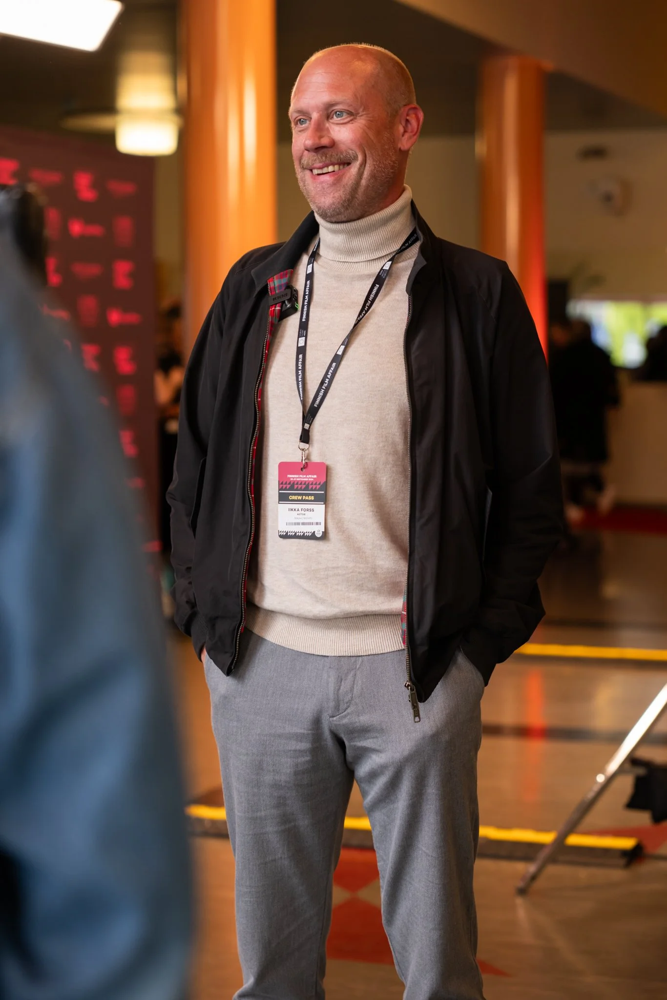 A smiling man with a crew pass around his neck, wearing a beige turtleneck and gray pants, standing indoors at a professional event.
