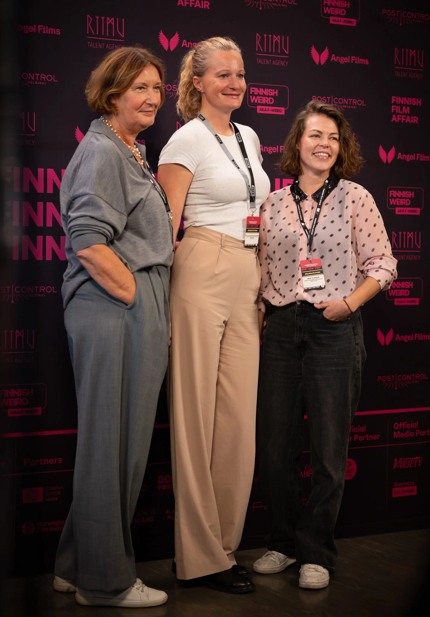 Three women standing together in front of a black backdrop with pink text and logos, smiling for a photo.