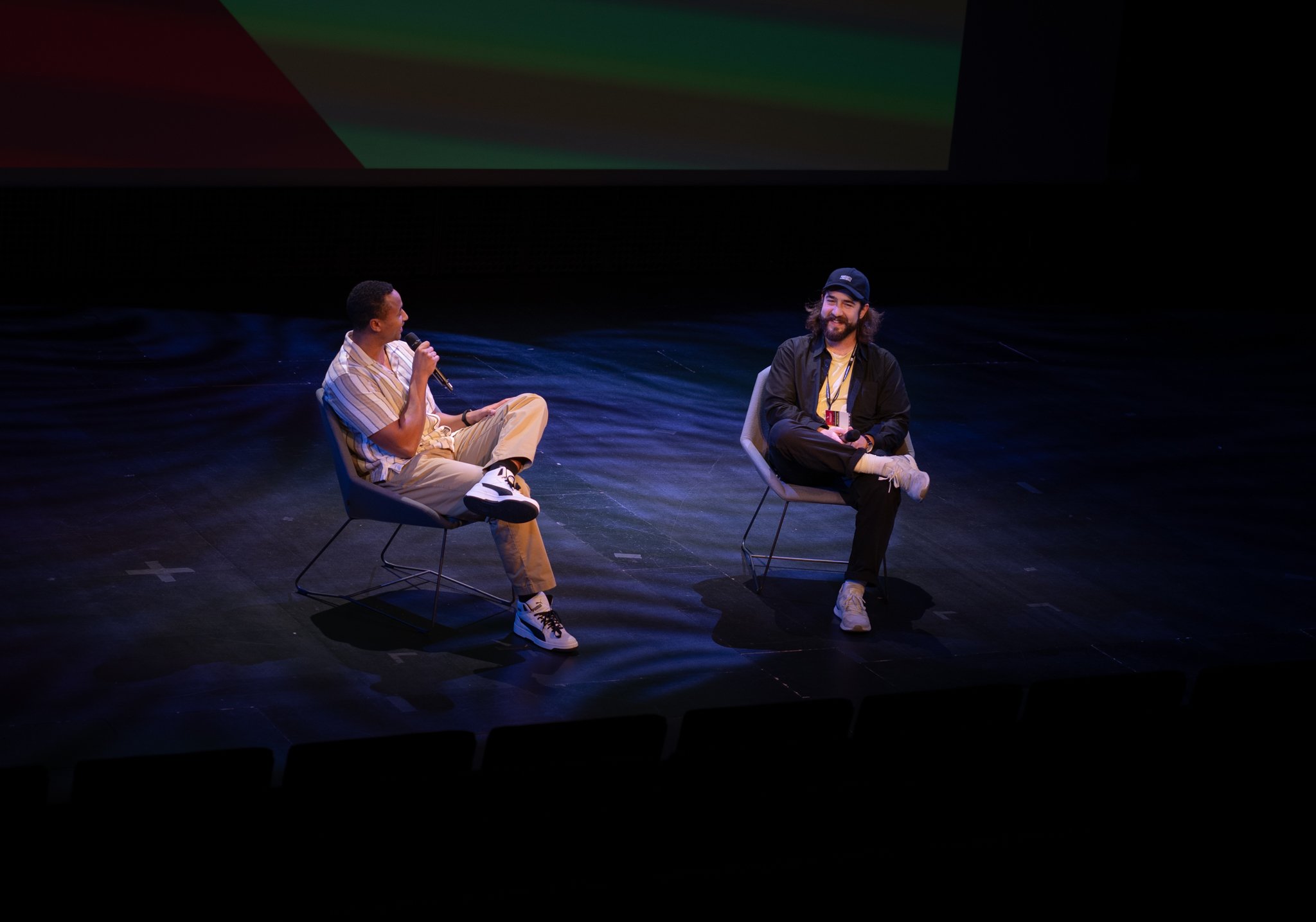 Two men sitting on chairs on a stage, engaging in a discussion, one holding a microphone and the other smiling at him. The stage has a dark floor and a large screen above them.