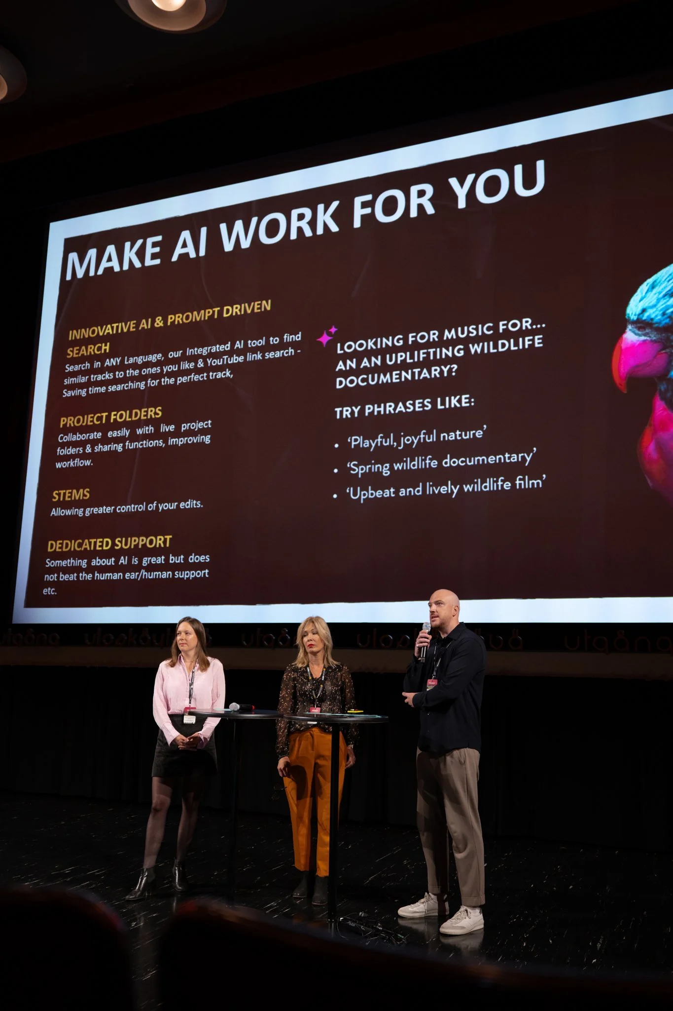 A presentation slide titled 'Make AI Work for You' is displayed on a screen behind three speakers standing on stage. The slide discusses innovative AI search, project folders, stems, and dedicated support, and offers suggestions for using AI to find 