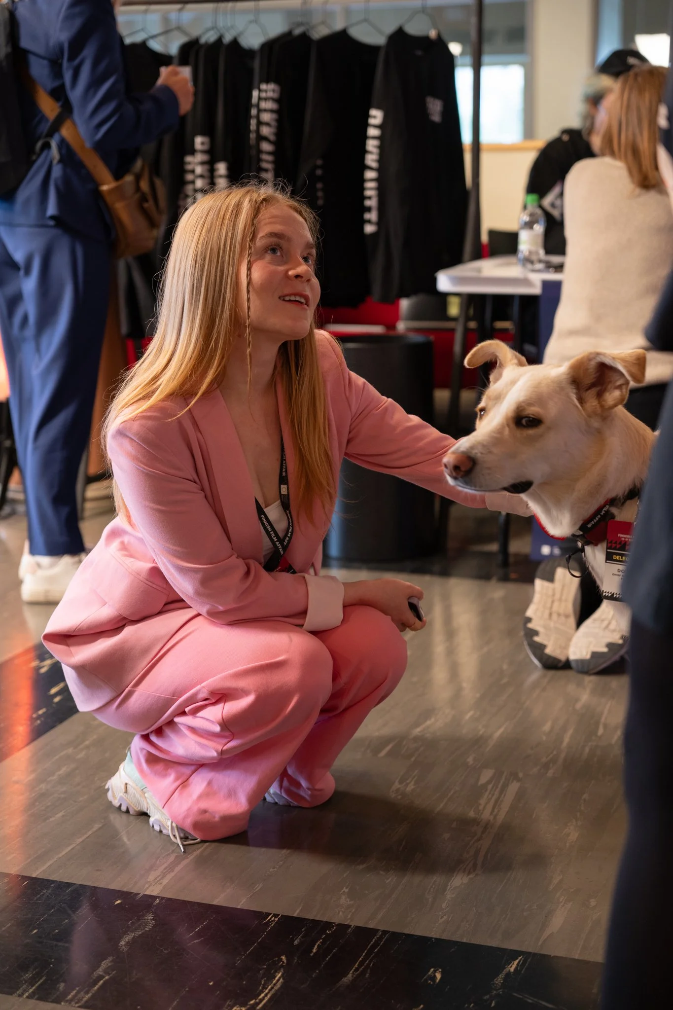 A woman in a pink jumpsuit squats on a dark wooden floor, petting a light-colored dog with a red collar. In the background, there are people, clothing racks with black jackets, and a table with drinks.