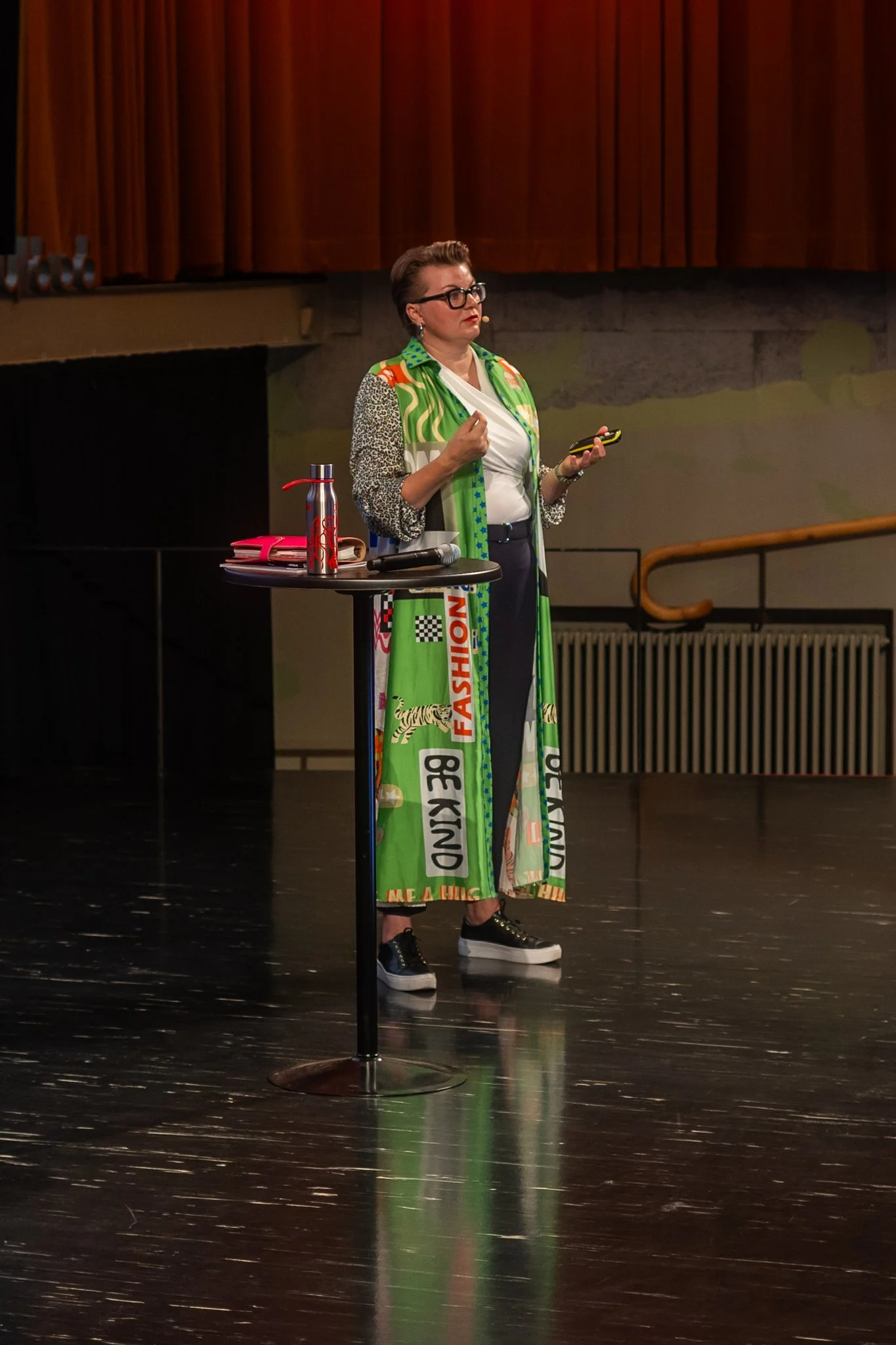 A woman with glasses and short hair standing on a stage, holding a phone, with a table beside her that has a water bottle, a notebook, and a microphone. She is dressed in a colorful, patterned outfit and sneakers, with a brown curtain in the backgrou