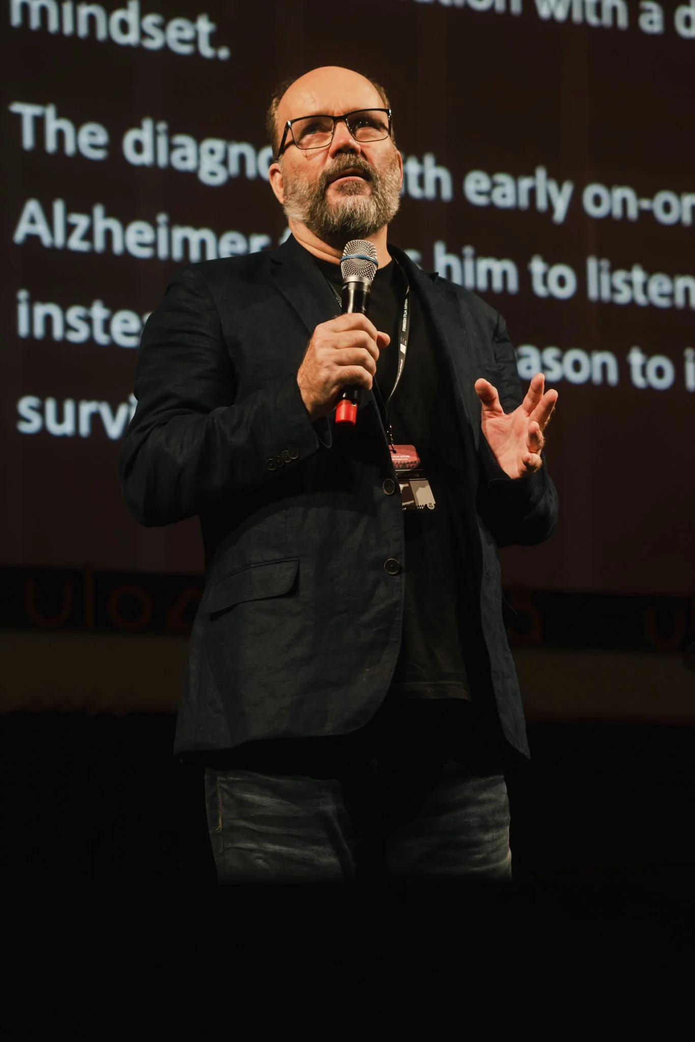 A man with glasses, a beard, and a bald head speaking into a microphone during a presentation or conference, with a large screen displaying text behind him.