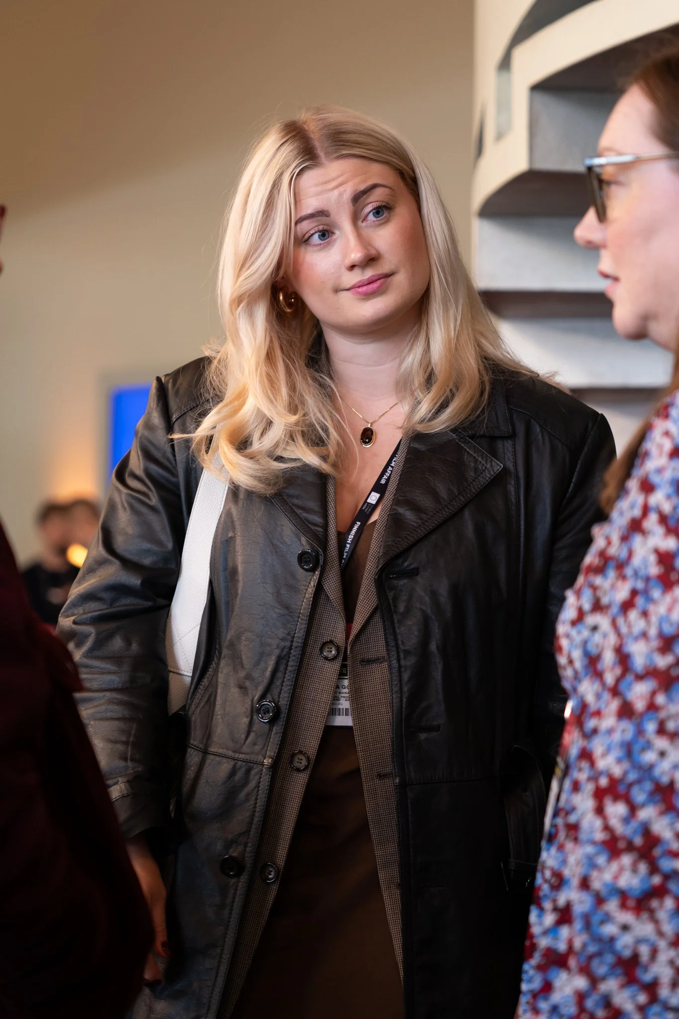 A woman with blonde hair wearing a black leather jacket and a necklace, looking with a skeptical or questioning expression at another woman with glasses and a colorful patterned shirt.