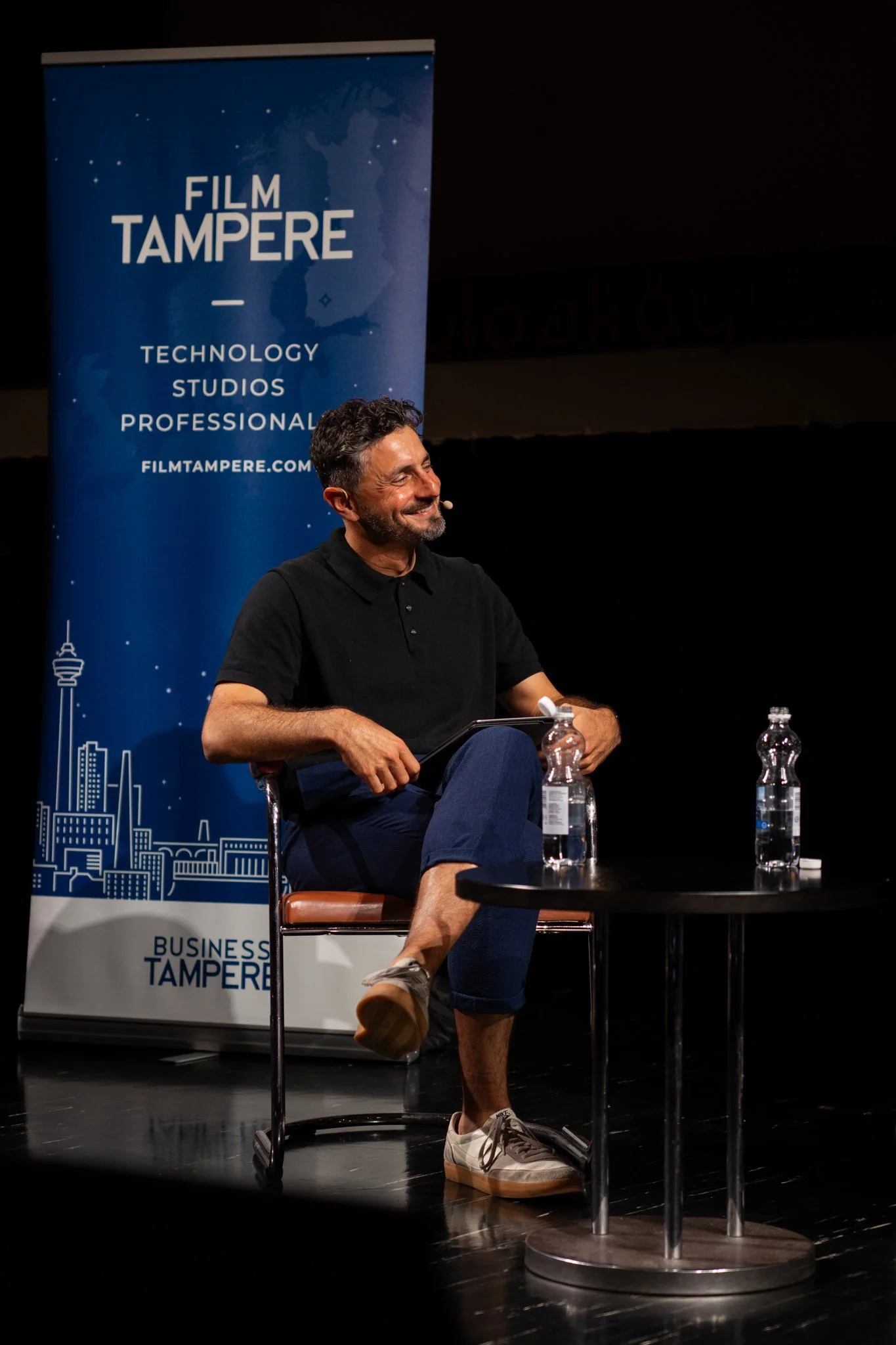 A man with dark hair and a beard, wearing a black polo shirt and jeans with rolled-up cuffs, sitting on a chair on stage during an event. He is smiling and looking to his left. On a small round table in front of him, there are two bottles of water. I