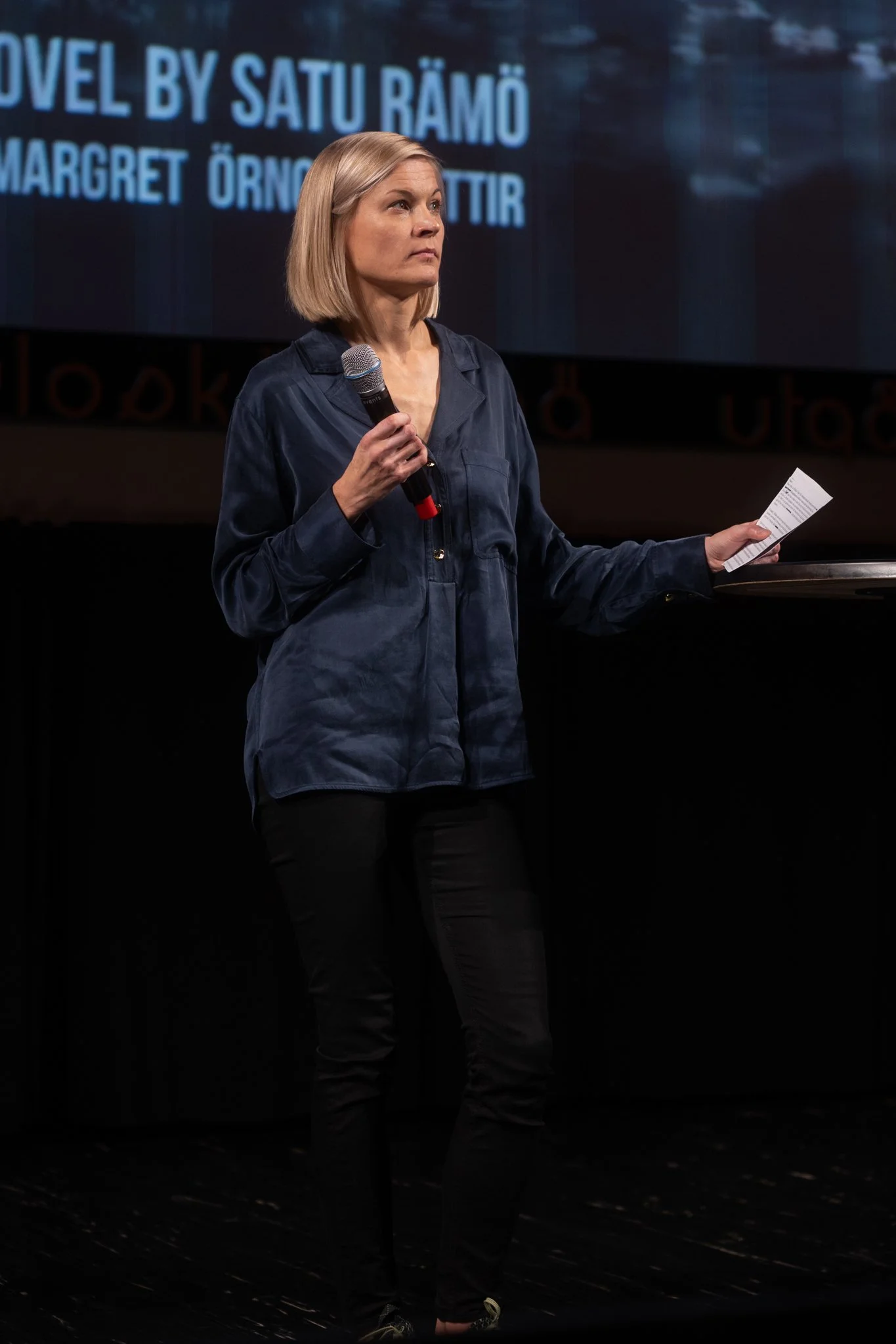 A woman with blonde hair holding a microphone and a piece of paper, standing on stage.