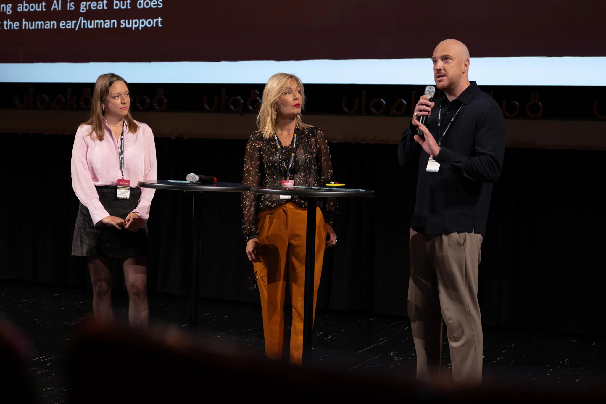 Three people standing on a stage participating in a discussion. One man is speaking into a microphone while two women listen. There is a small table with a microphone and other items in front of them. A large screen with text is in the background.