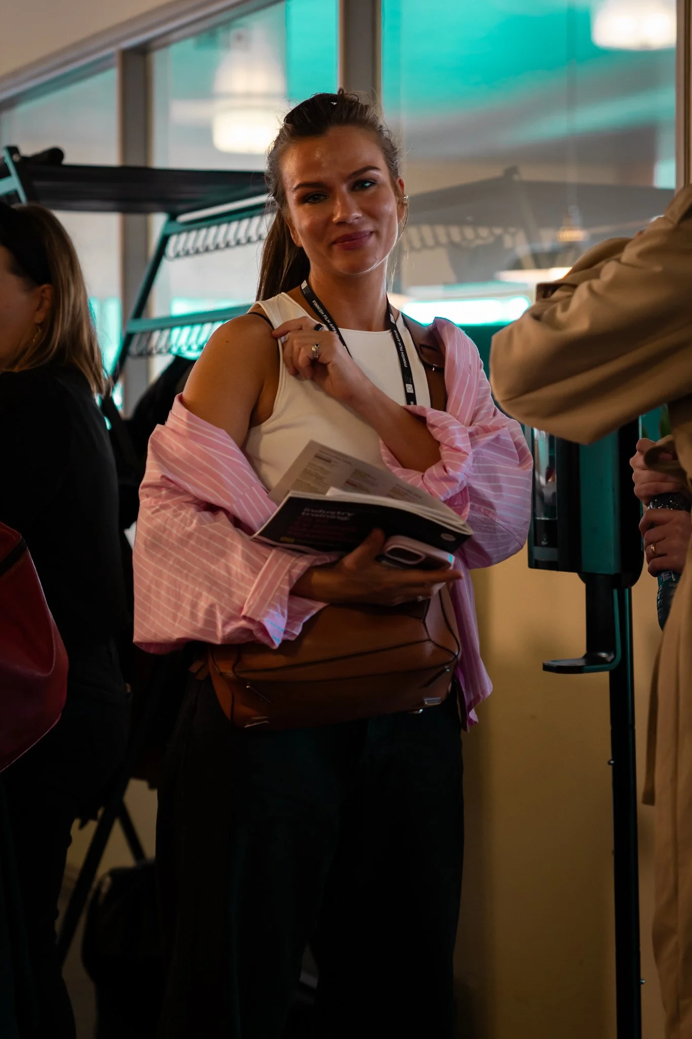 A woman standing in line at an airport, holding a pamphlet and a brown leather purse, wearing a white tank top with a pink striped shirt draped over her shoulders, and a black lanyard around her neck.