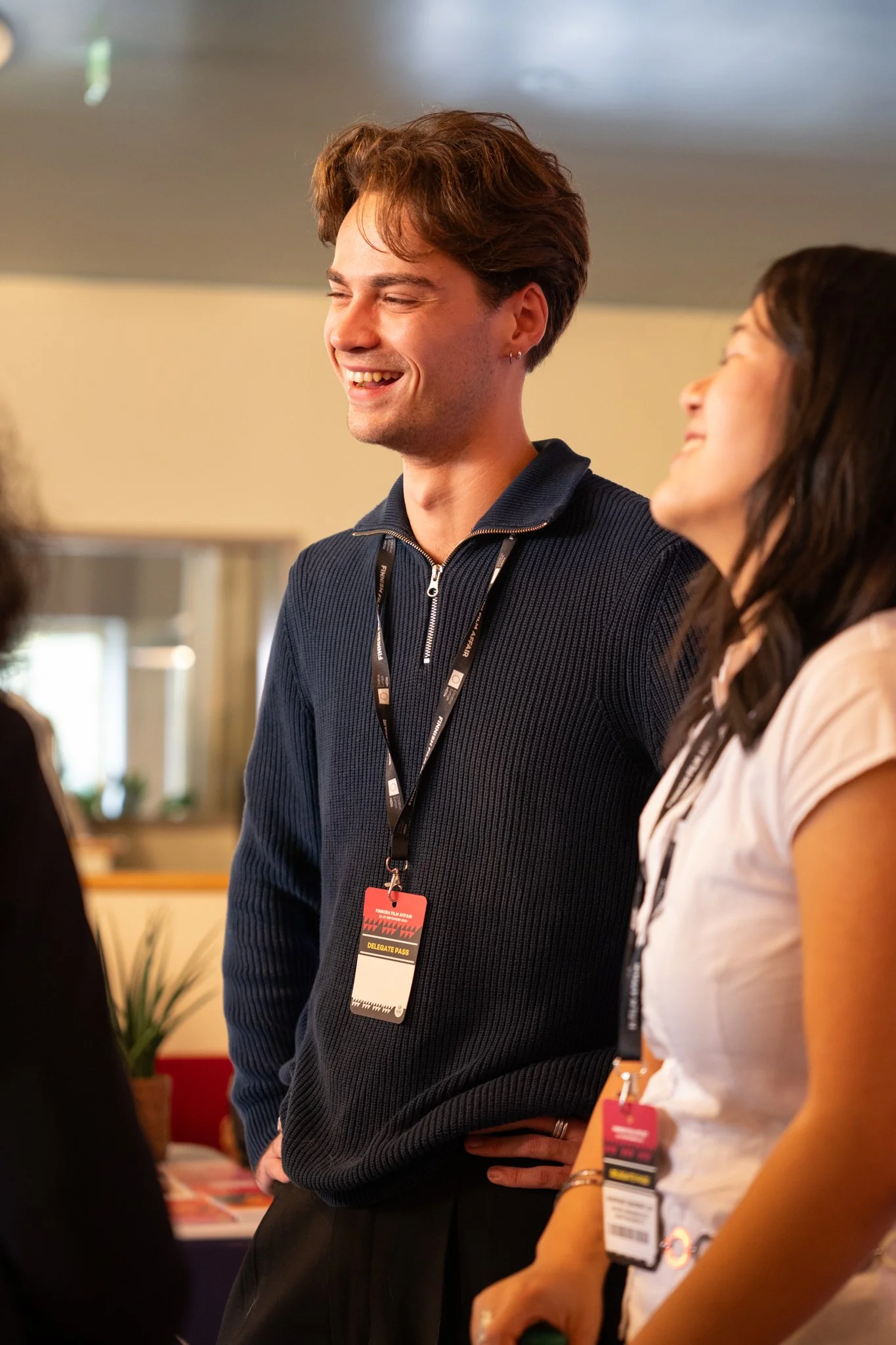 A young man and woman smiling and engaging in conversation at an indoor event, wearing conference badges around their necks.
