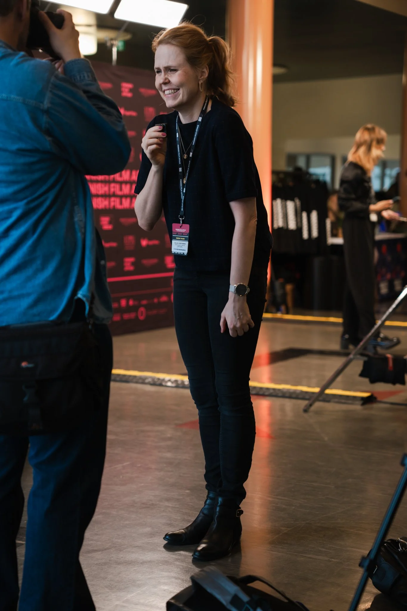 A woman with red hair smiling and talking to a man, wearing a black outfit and a lanyard, at an indoor event or conference.