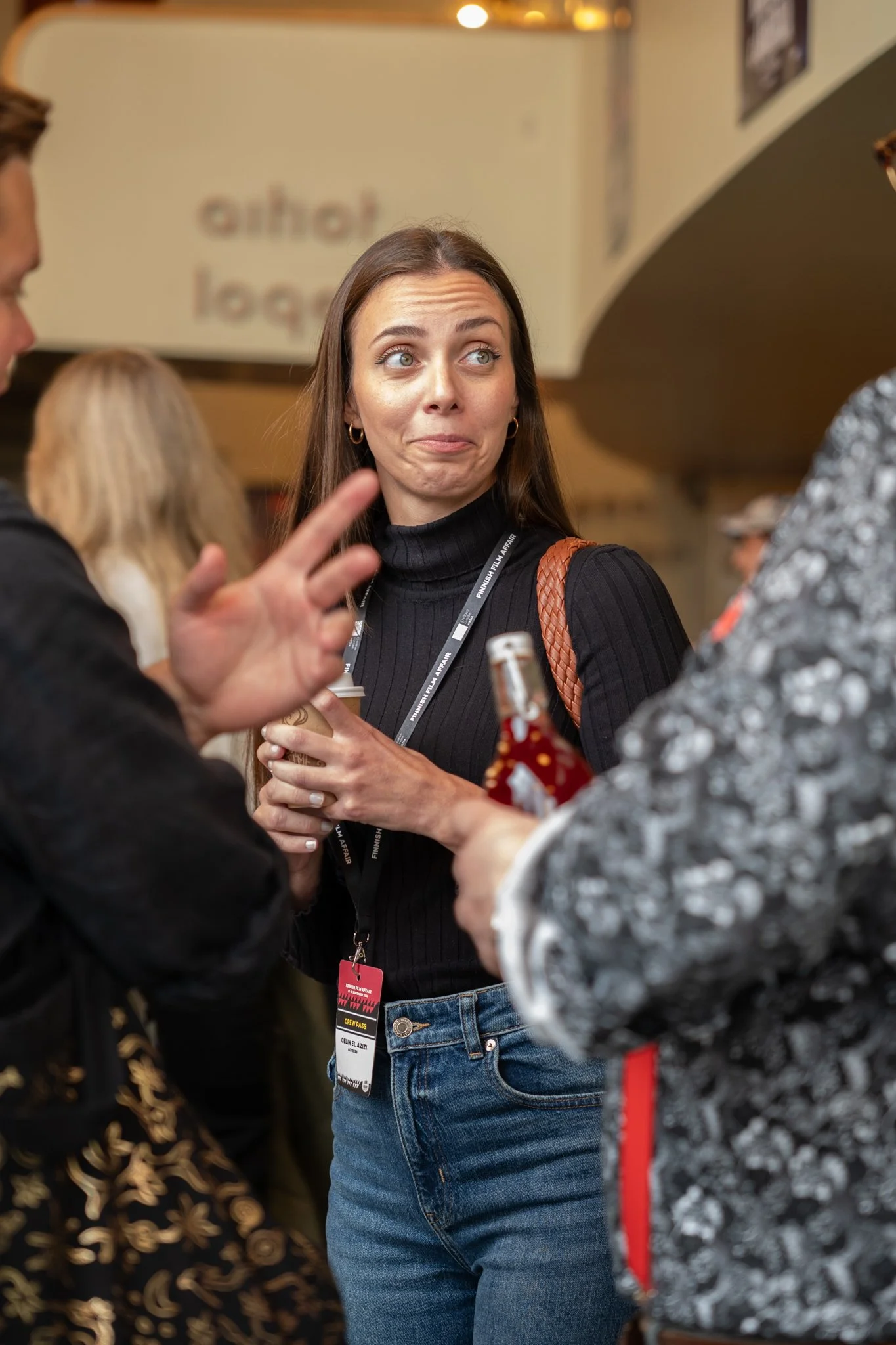 A woman with long brown hair, wearing a black turtleneck and blue jeans, holds a coffee cup and appears to be listening while talking to two other people at a social event.
