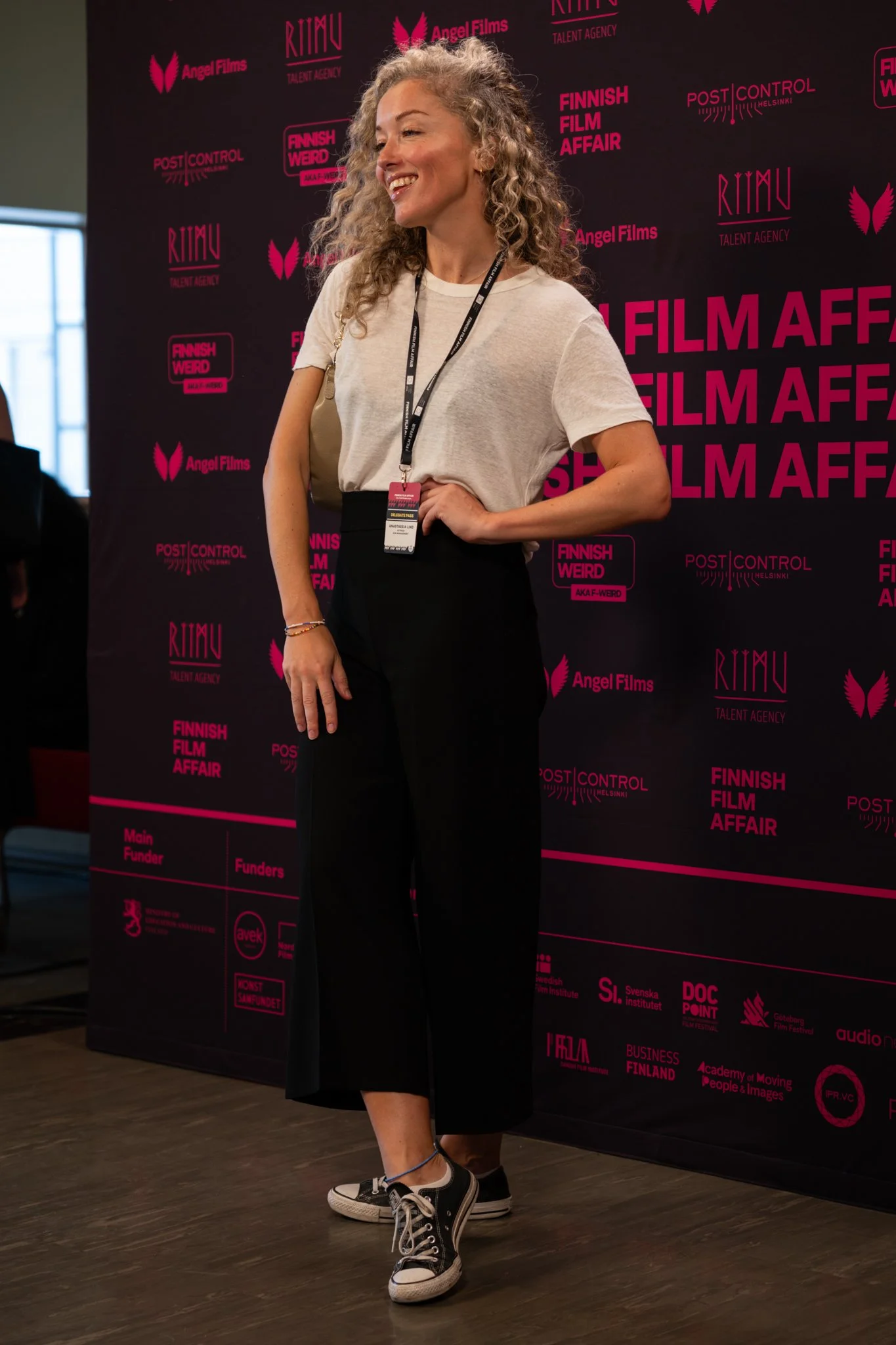 A woman with curly blonde hair wearing a casual white t-shirt, black pants, and black sneakers, standing in front of a red and black FILM AFFAIR backdrop at a film festival event, smiling and posing.