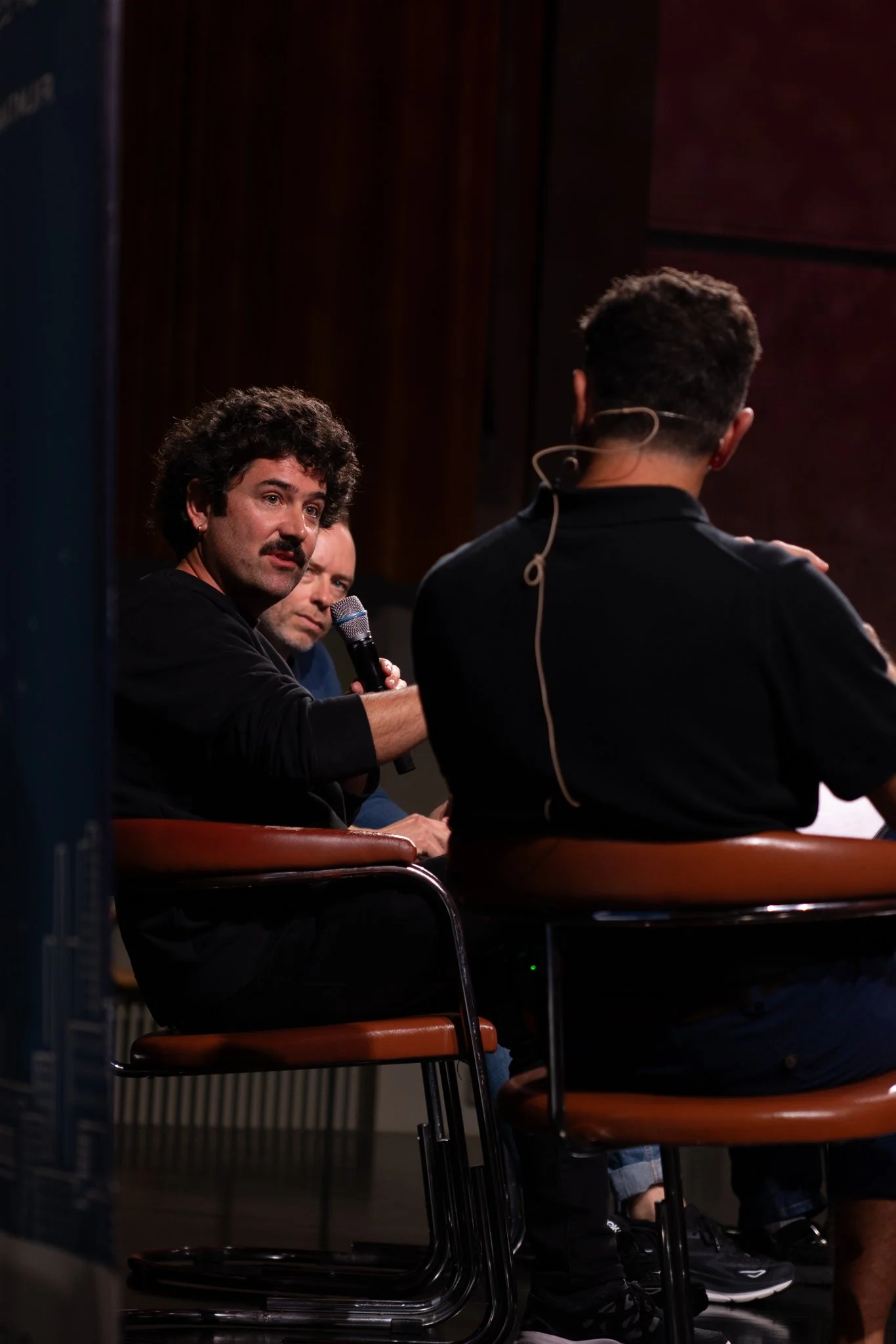 Three men sitting on chairs in a dark room, one of them with a microphone, engaging in a discussion or panel.