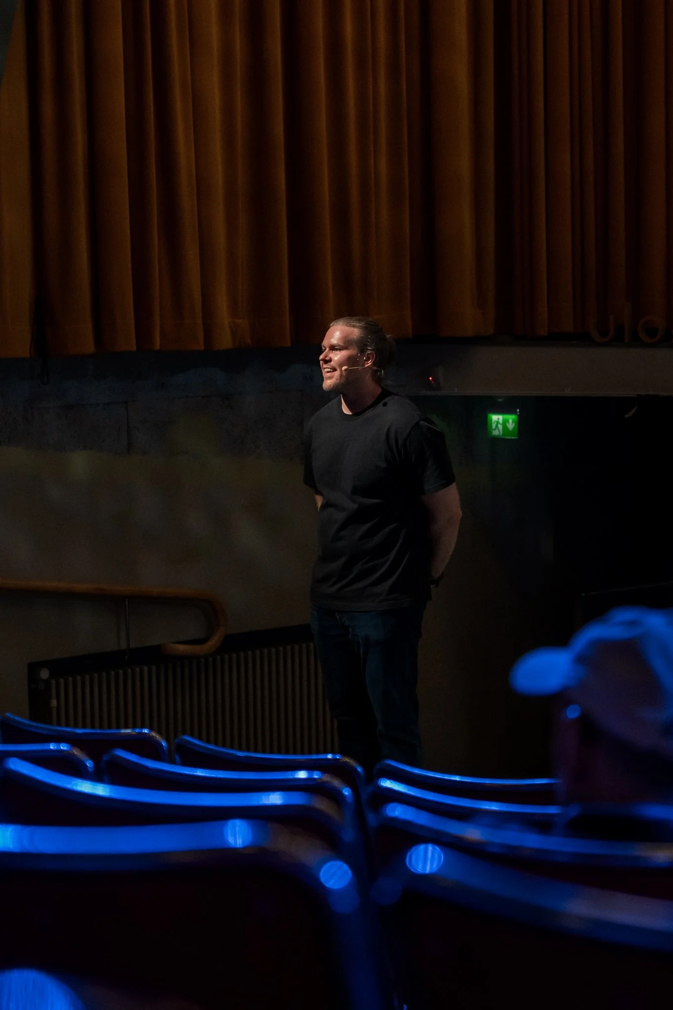 A man standing on a stage in front of a brown curtain, smiling and wearing a black t-shirt, with blue theater seats in the foreground.