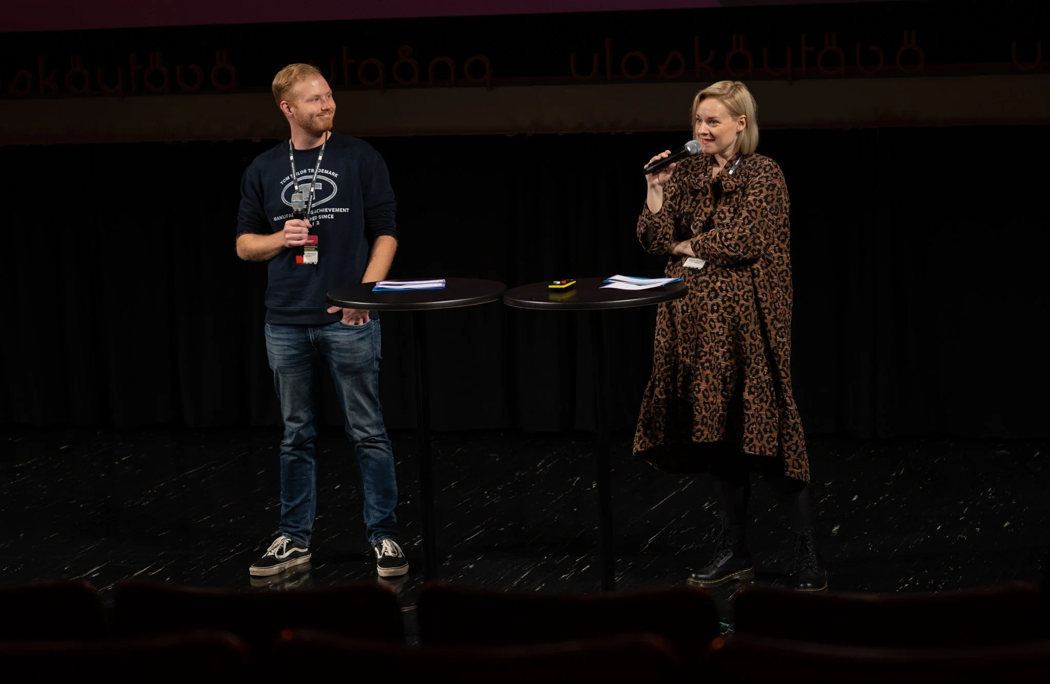 A woman and a man standing on a stage with black curtains, engaged in a discussion. The woman is speaking into a microphone, wearing a leopard print dress, while the man is holding a microphone, dressed in jeans and a sweatshirt. Two small round tabl